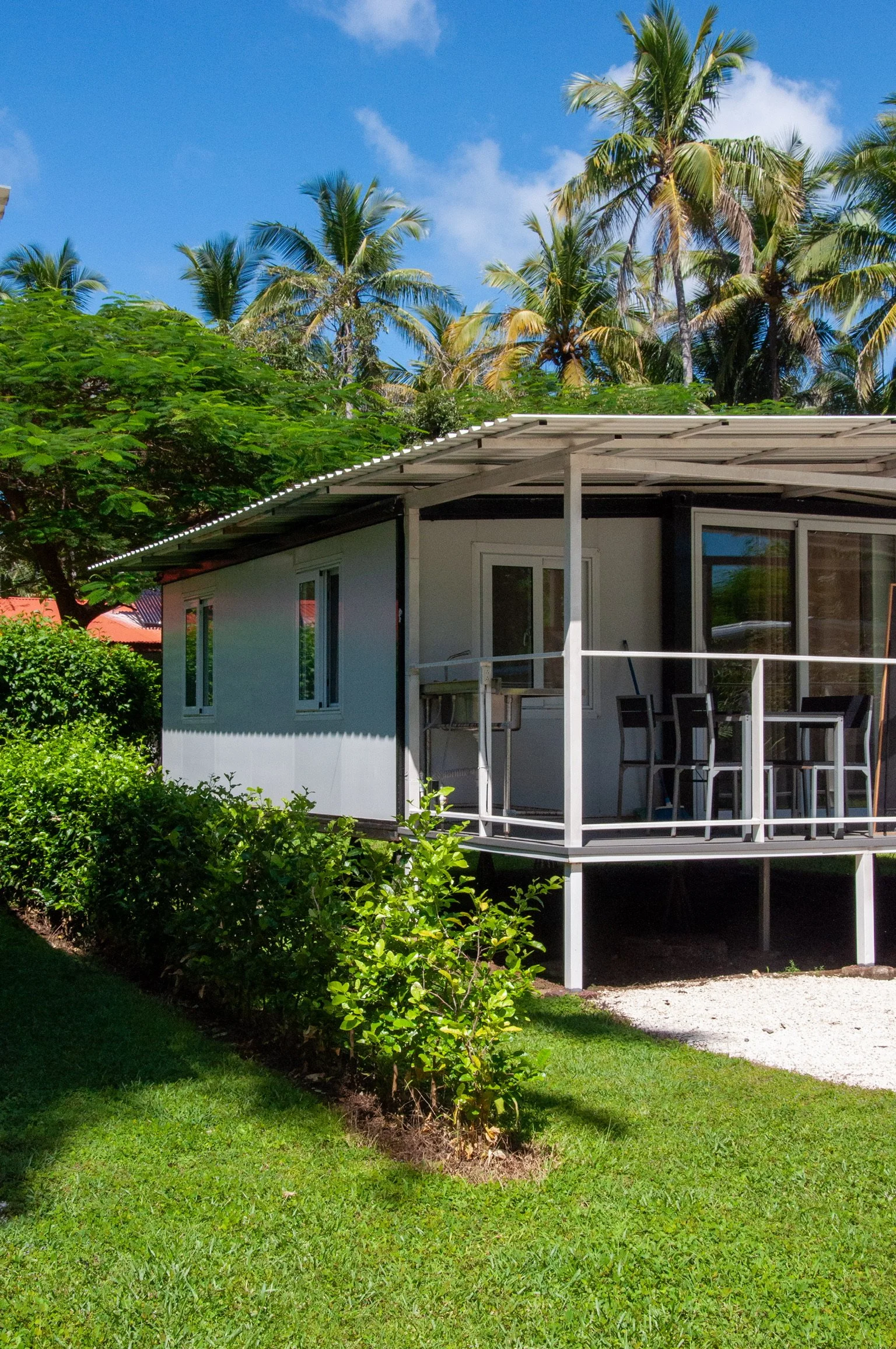 A modern house with a balcony, surrounded by lush green plants and tall palm trees under a bright blue sky.