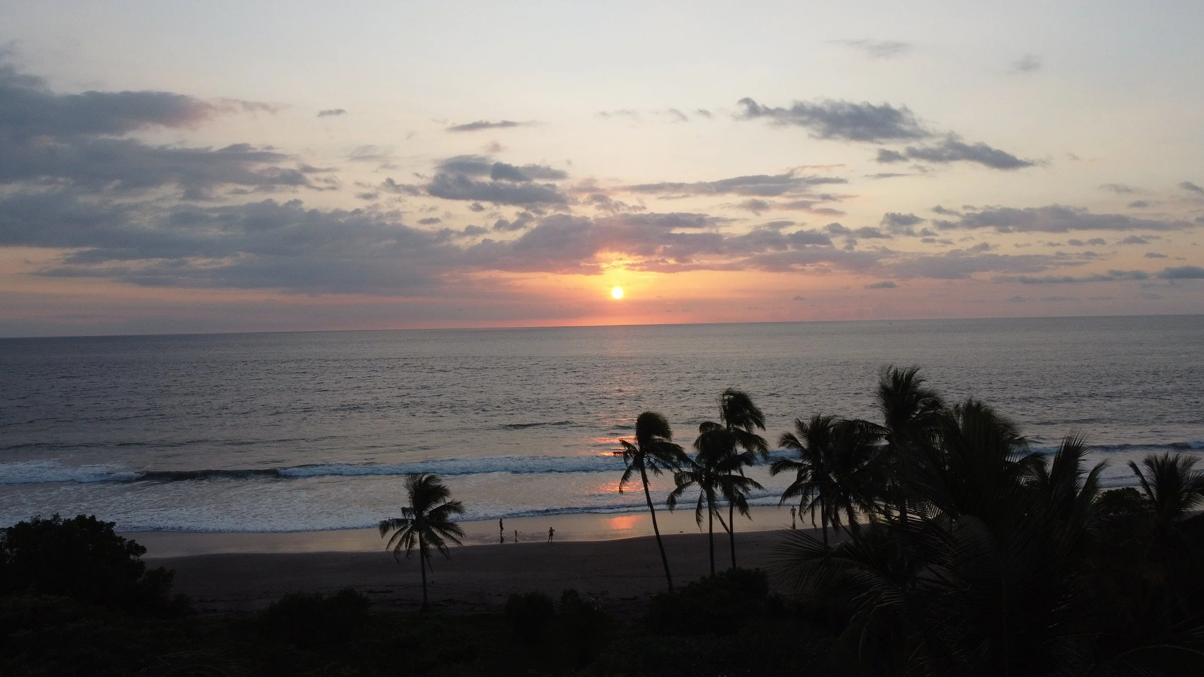 A sunset over the ocean with Lava rocks in the foreground and waves crashing against them.