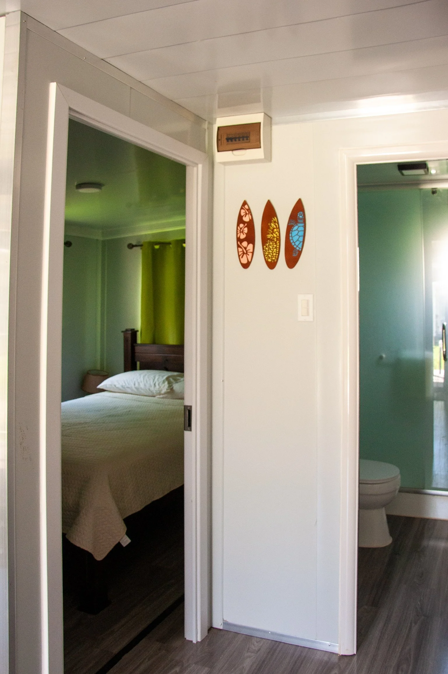 Bedroom visible through a sliding door with a bed, beige quilt, and a nightstand with a lamp. On the wall adjacent to the door are three decorative wooden leaf-shaped art pieces. A bathroom with a toilet and a window is visible through a mirror reflection on the right.