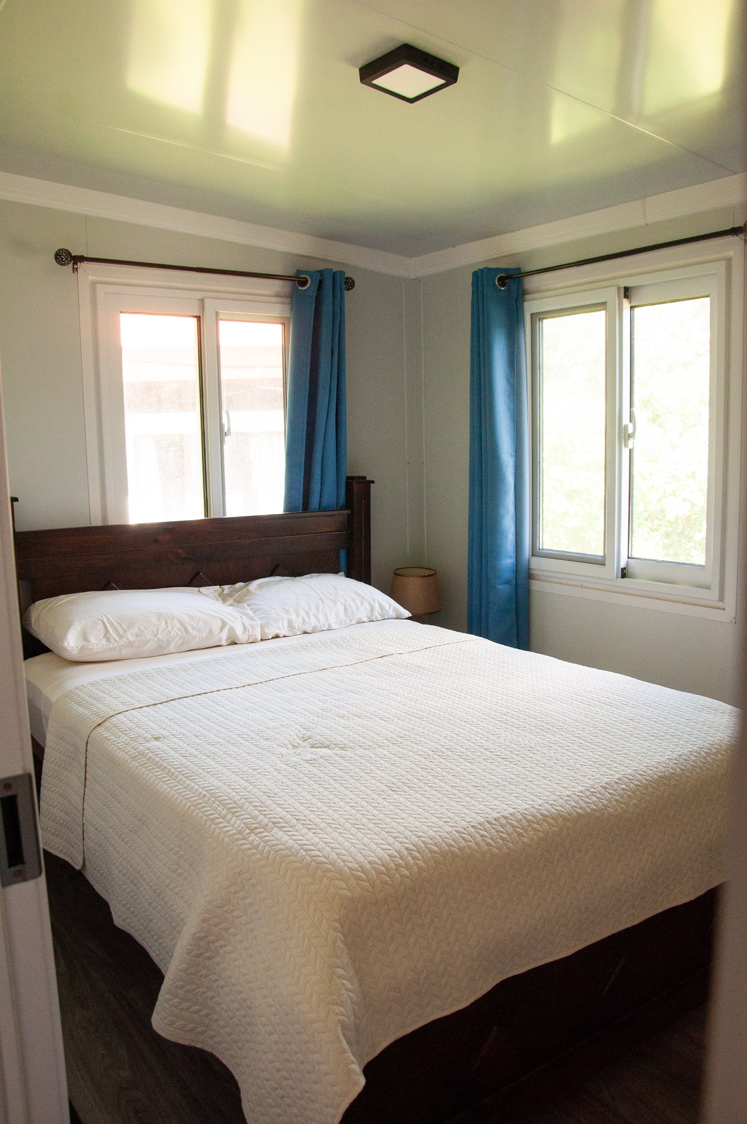 A bedroom with a bed covered in a white quilt and two pillows. There are two windows with blue curtains and a small wooden nightstand with a lamp. The ceiling has a square modern light fixture.