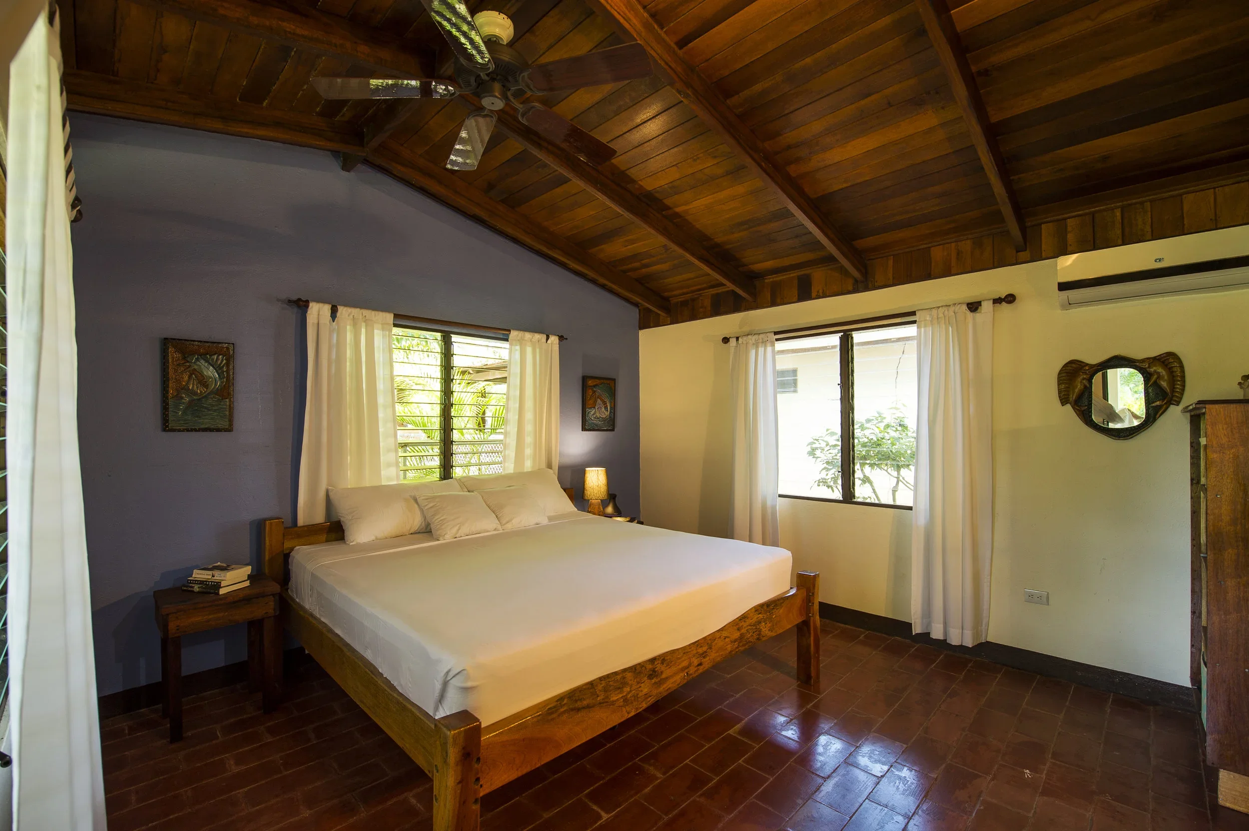 A bedroom with a wooden vaulted ceiling, a bed with white linens, two windows with white curtains, a nightstand with books and a lamp, and decorative artwork on the walls.
