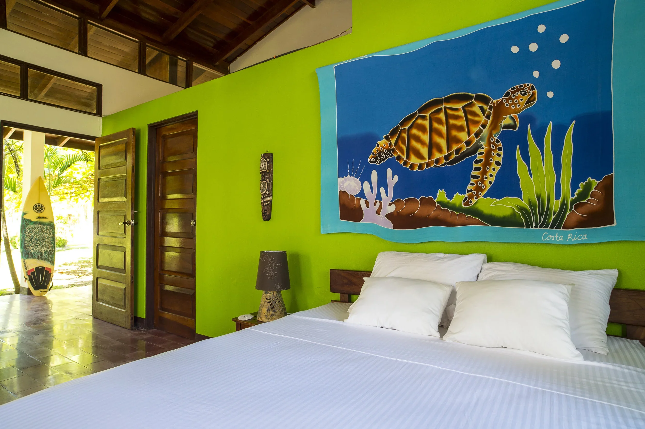 Interior of a bright room with green walls and a large bed with white bedding. A colorful painting of a turtle marked Costa Rica hangs on the wall. There are wooden doors, a black lamp on a side table, and a surfboard propped against the wall near the open door.