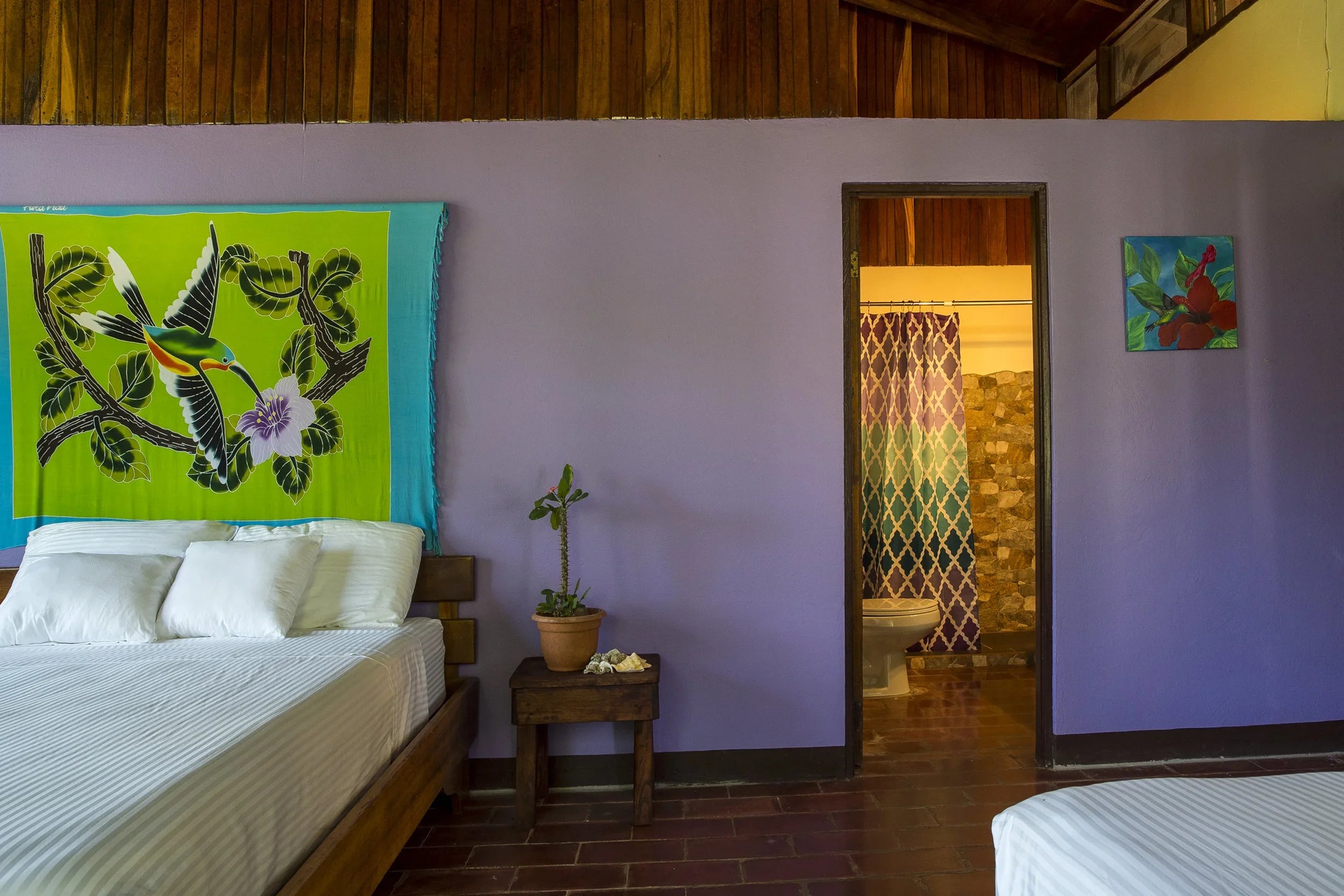A bedroom with purple walls, a large bed with white striped bedding, a wooden nightstand with a potted plant, and a colorful wall hanging of a hummingbird and flower. An open door reveals a bathroom with patterned shower curtain and stone wall.