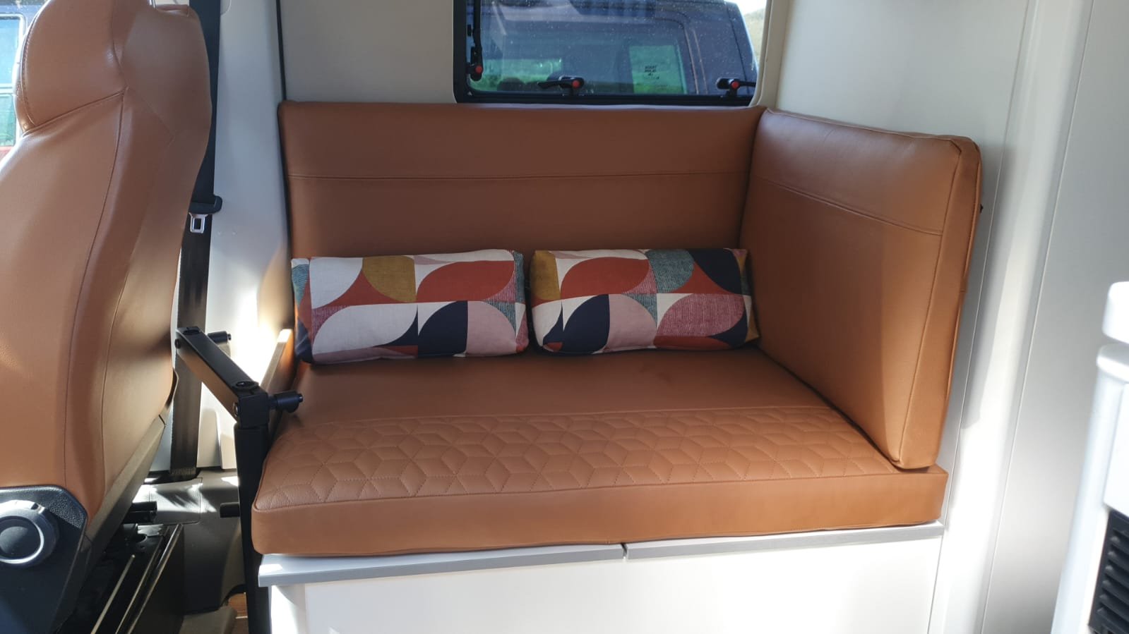 Inside a vehicle, a tan leather bench seat with two patterned pillows and a window showing a vehicle outside.