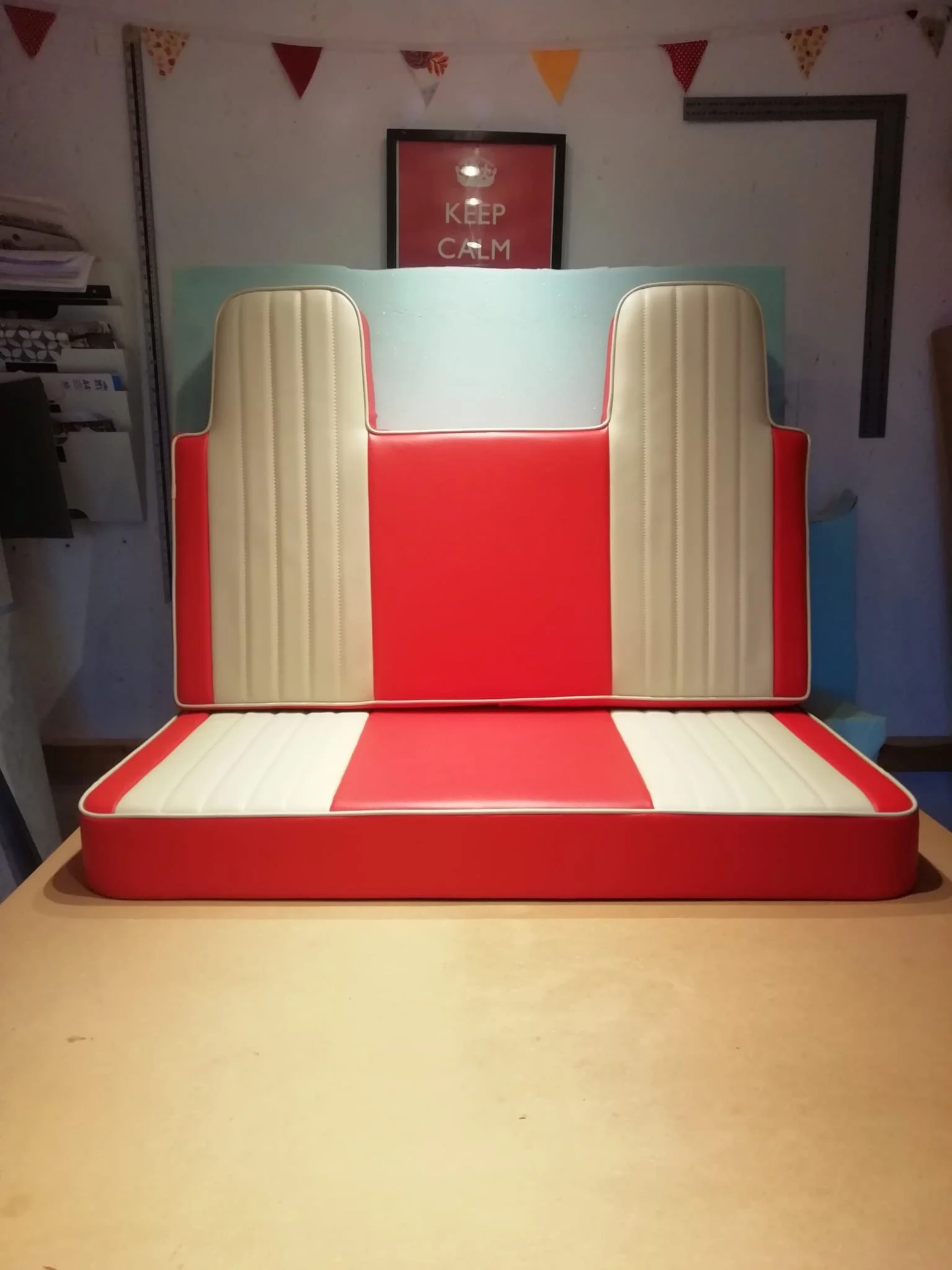 Red, white, and blue padded crib with a backrest, placed on a wooden table, with a 'Keep Calm' poster and colorful bunting in the background.