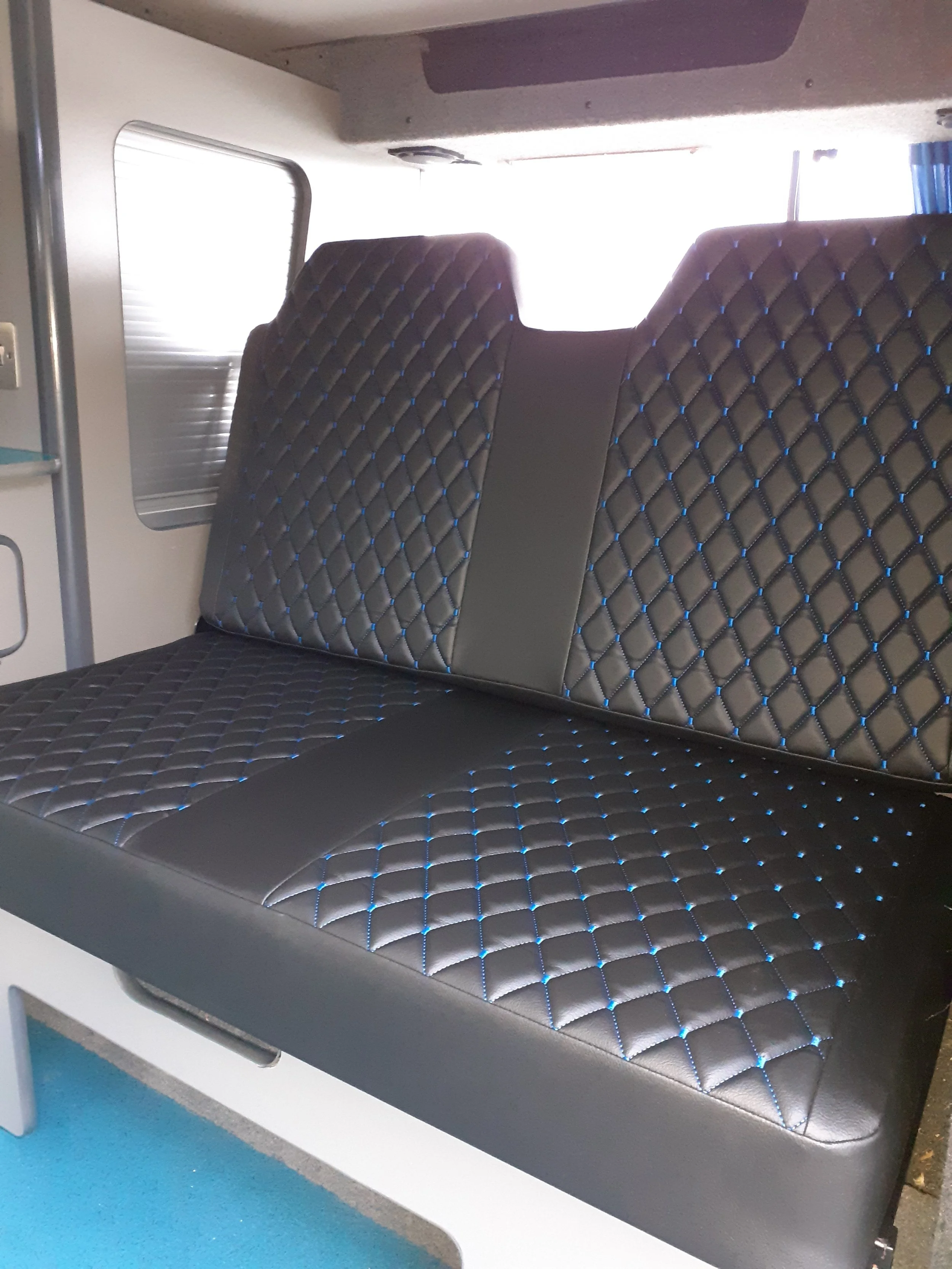 View of a train or bus interior with black padded seats that have a blue diamond stitching pattern, a window, and blue flooring.
