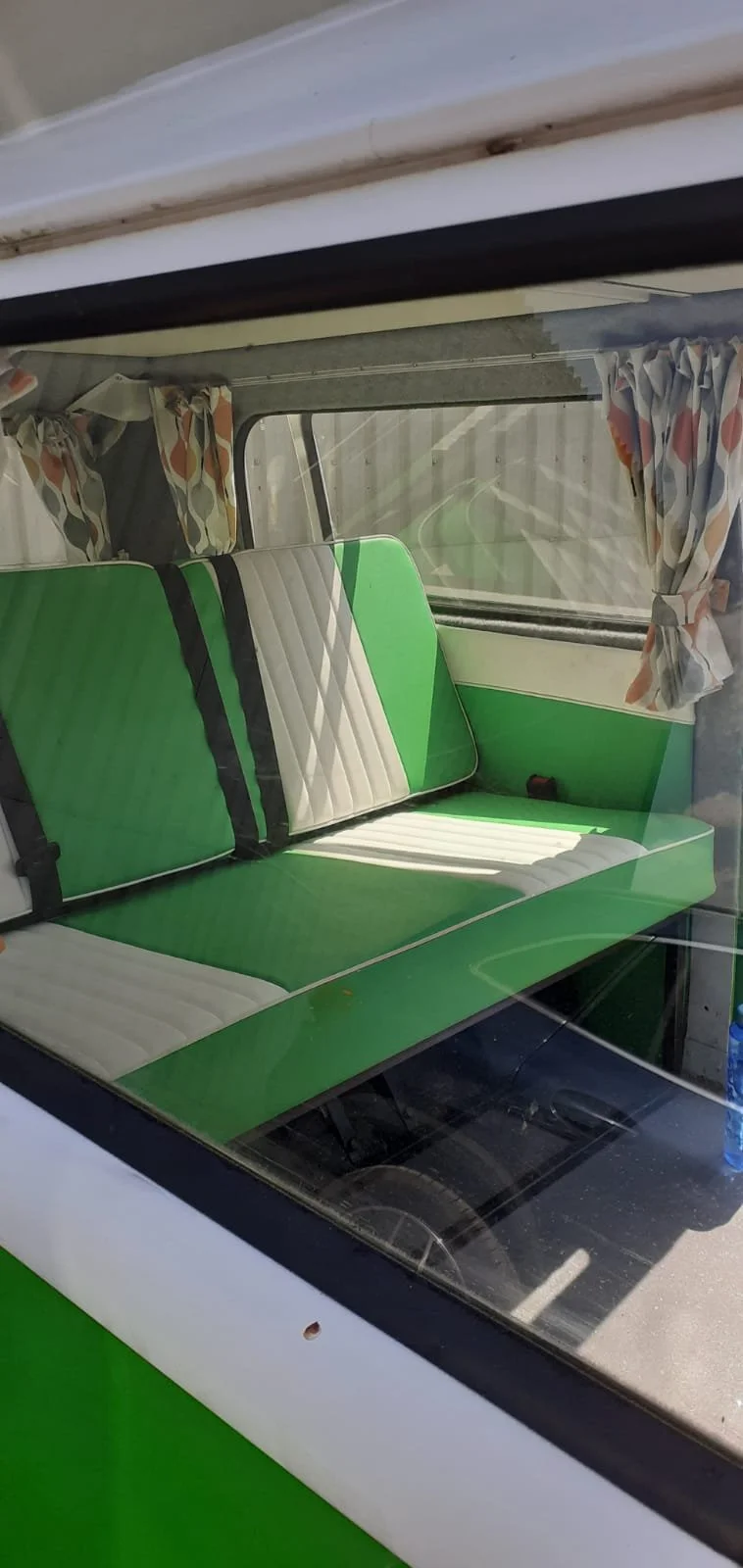 Inside view of a vintage green and white camper van with a bench seat, window with floral curtains, and a view of the road outside.