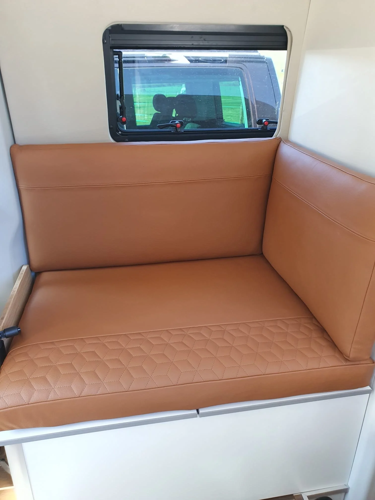Inside a camper or RV, showing a tan leather seat or sofa with a diamond quilted pattern on the cushion, and a window with a view of a green field outside.