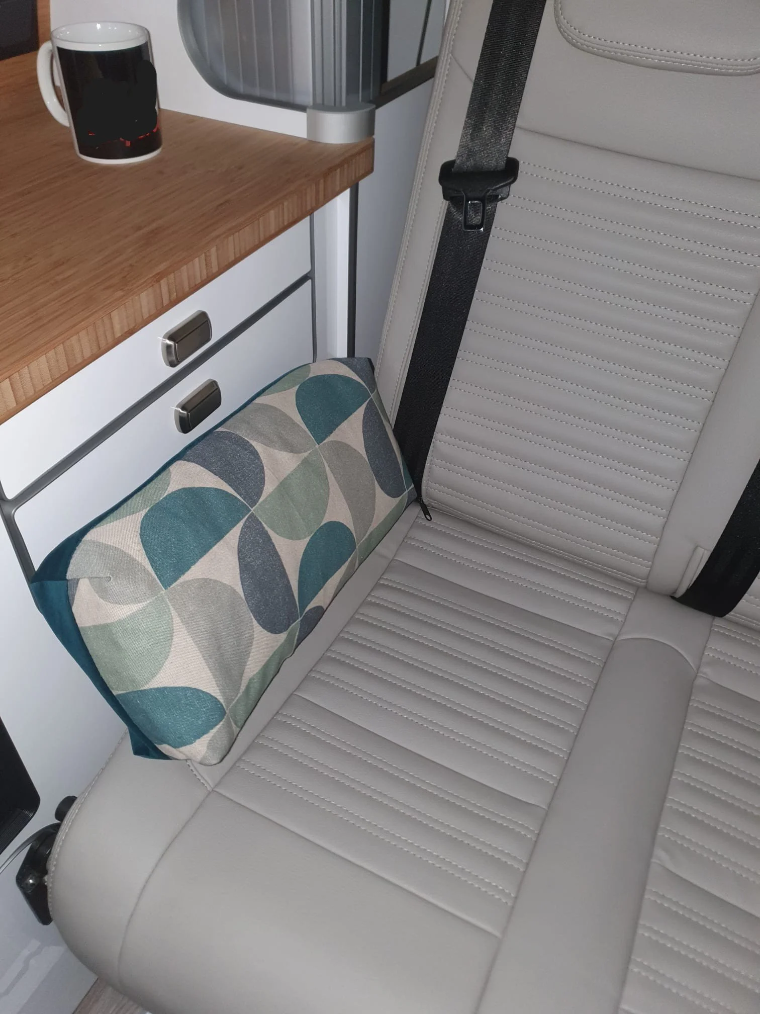 A white leather car seat with horizontal stitching, a black seatbelt, and an armrest, with a patterned pillow resting on the seat.