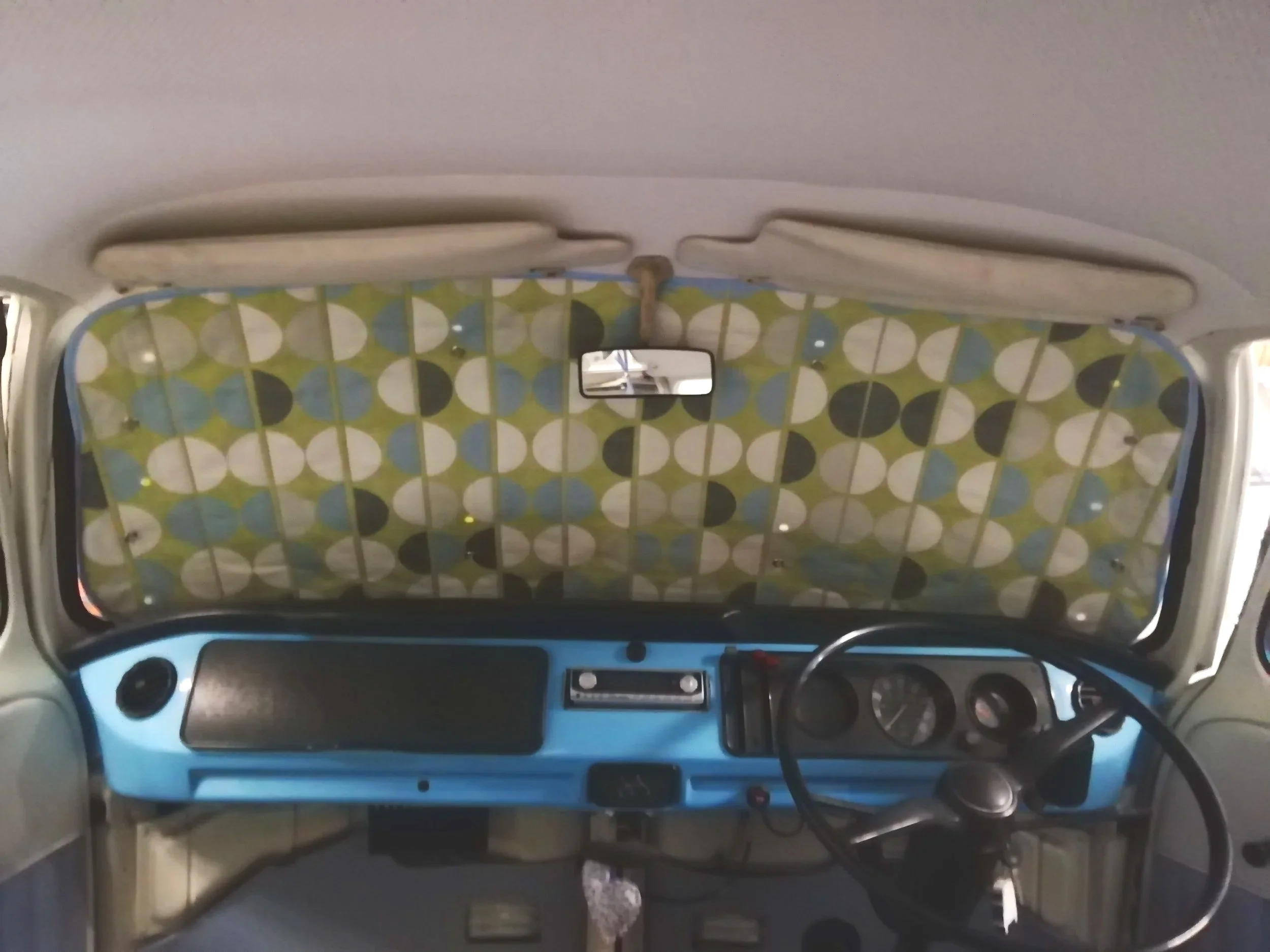 The interior of a vehicle featuring a dashboard with a radio, round air vents, and a small mirror on the windshield. The back of the vehicle shows a patterned divider with a repeating circle design in green, white, black, and blue.