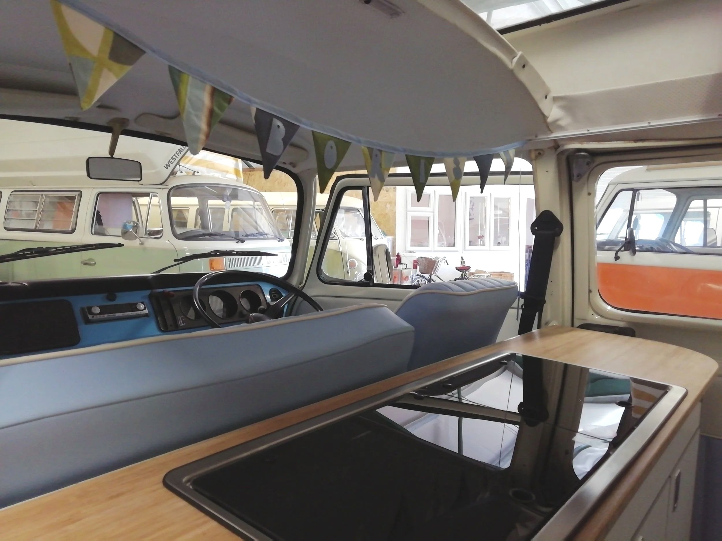 Interior of a vintage Volkswagen van with bunting decorations hanging from the ceiling, a dashboard with a radio and steering wheel, and a seat with a table in the foreground. Outside the window, other similar vans are visible, indicating a possible 