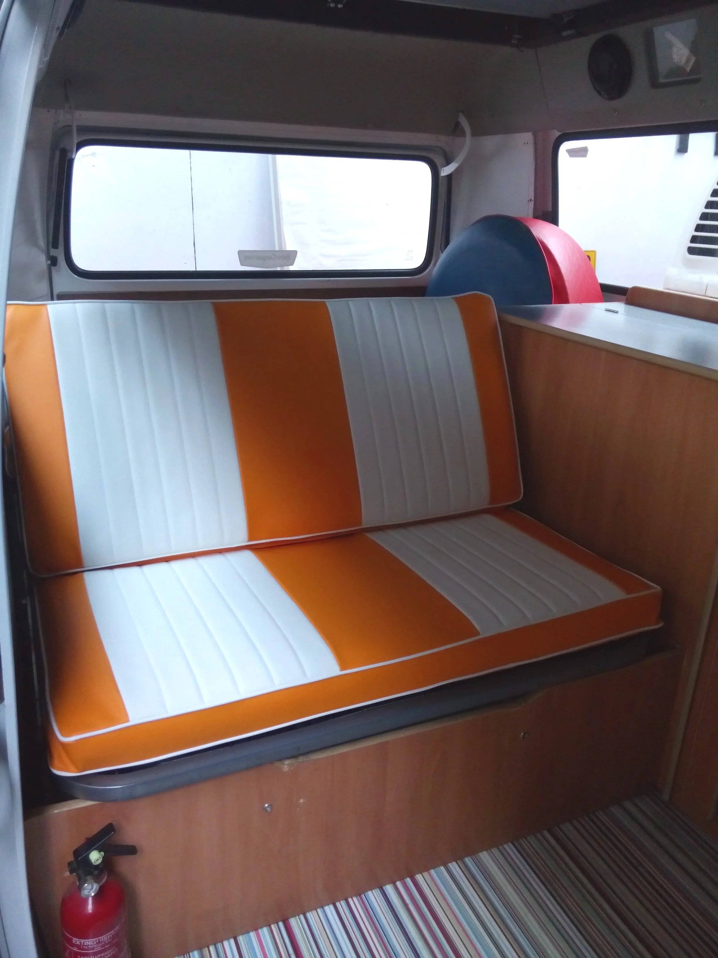 Interior of a vehicle with a striped orange and white cushioned bench seat, a wooden panel below, a fire extinguisher in the corner, a window with a reflection, a blue and red circular object, and a striped carpet floor.