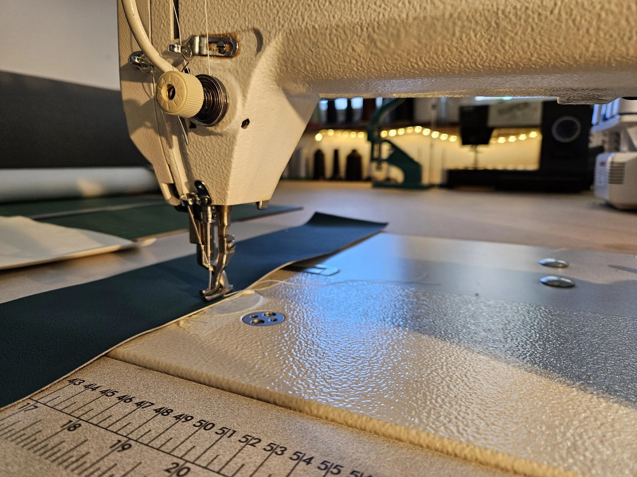 Close-up of a sewing machine needle, bobbin, and part of the sewing surface with measurement markings, in a cozy, well-lit room with string lights in the background.
