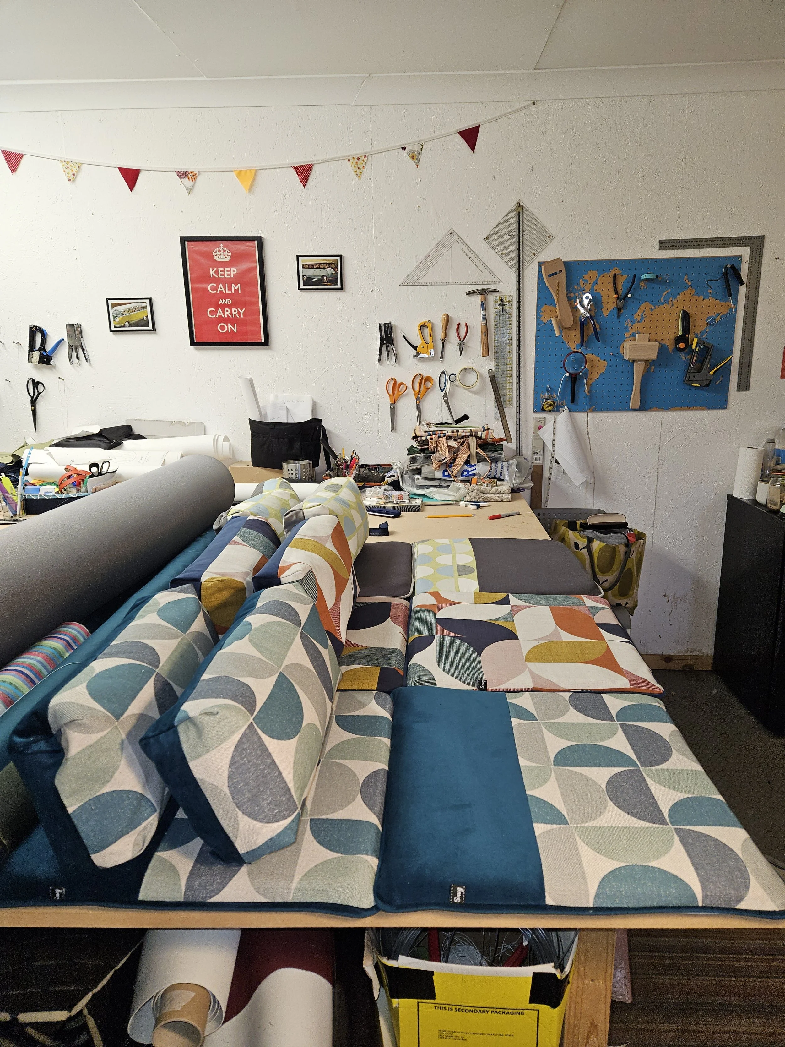 A cluttered workspace with a couch covered in colorful, geometric-patterned cushions. The wall behind features framed pictures, a motivational poster that says "Keep Calm and Carry On," and various measuring tools on a pegboard. There are rolls of pa
