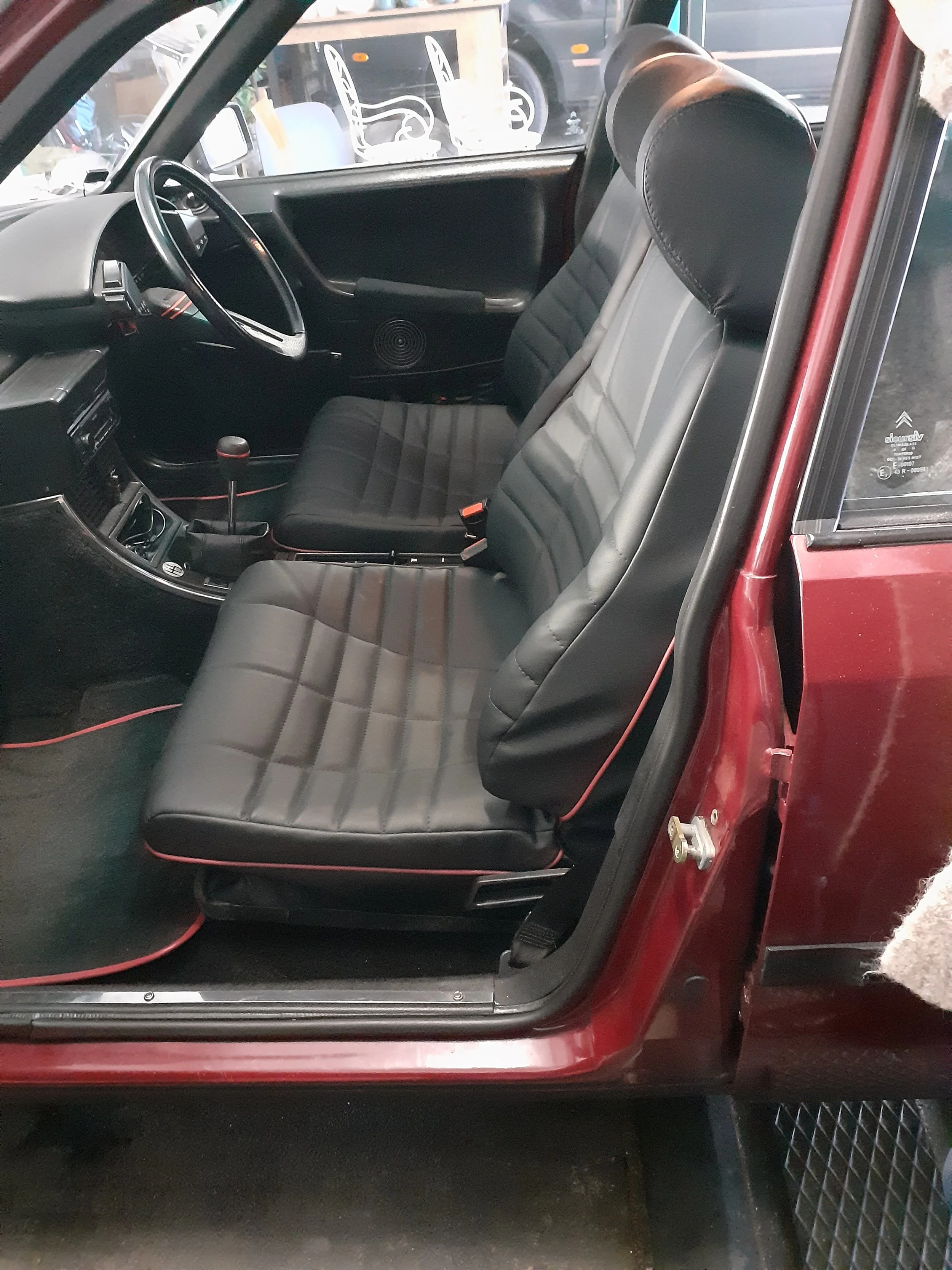 The interior of a vintage car showing the black leather front seats, steering wheel, dashboard, gear shift, and door panel. The car is parked indoors, and some chairs are visible outside through the window.