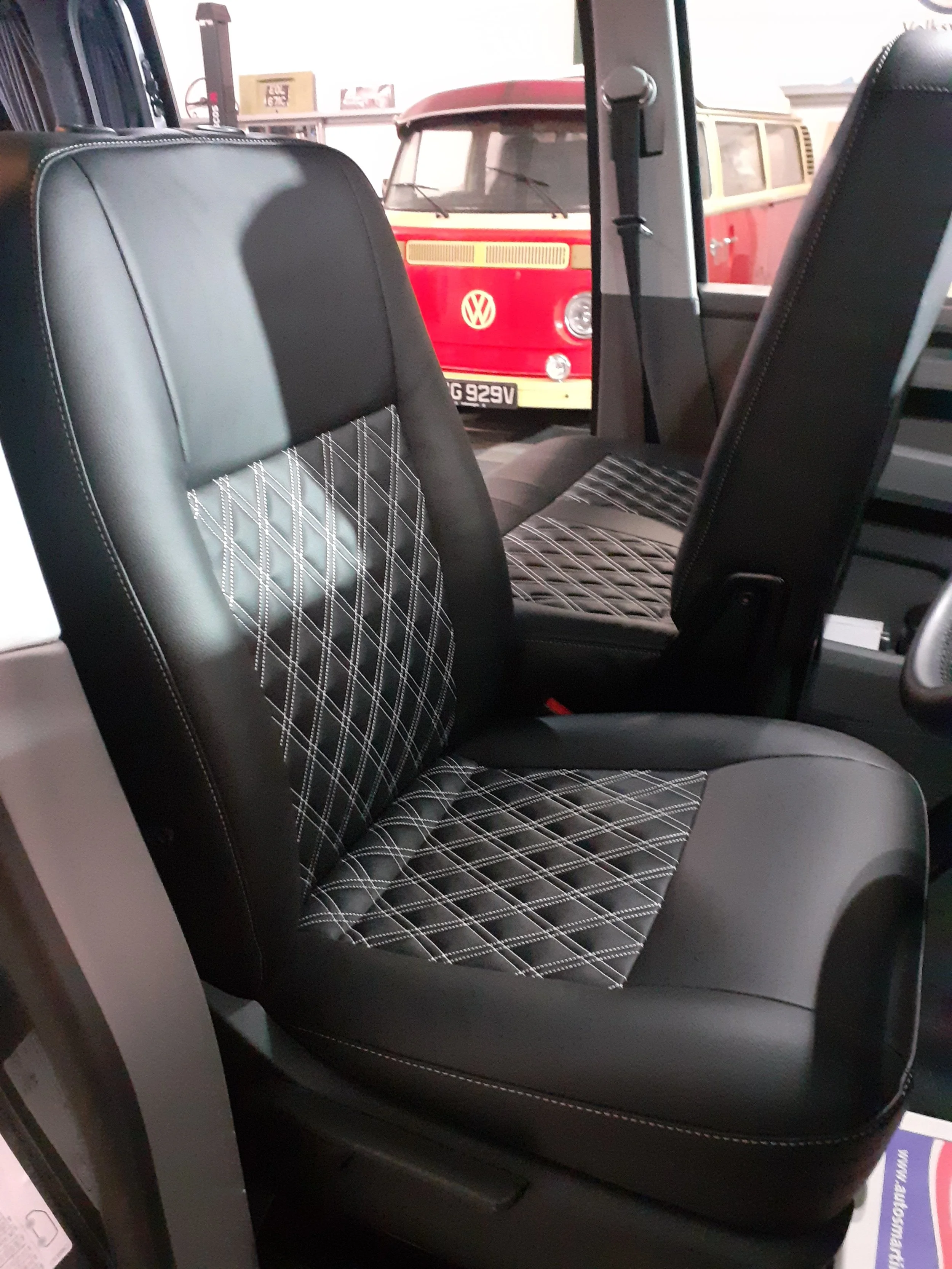 The image shows the interior of a vehicle, focusing on a black seat with a diamond-patterned fabric insert. In the background, there is a red and cream vintage Volkswagen van seen through the window.