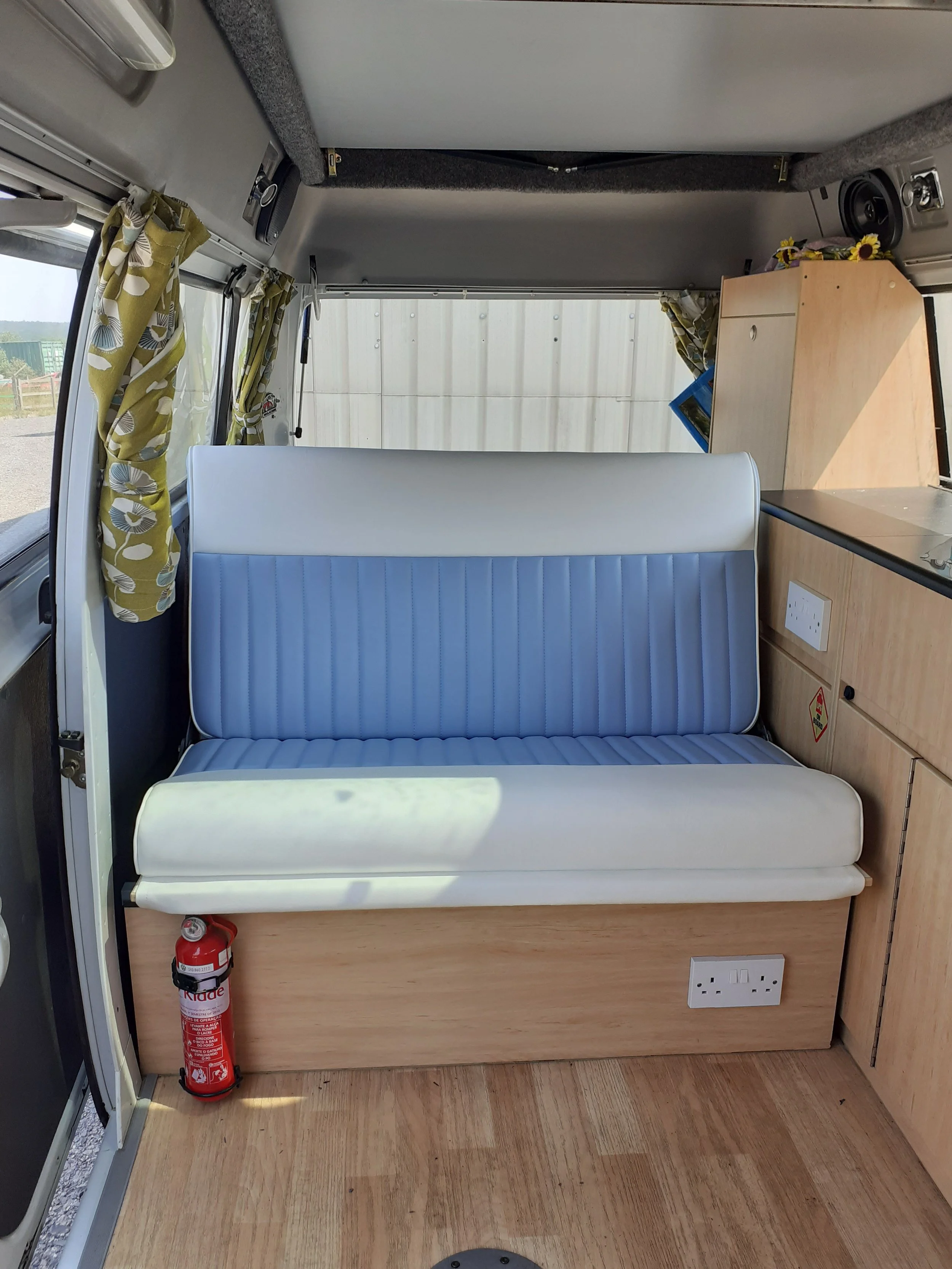 Interior of a campervan showing a bench seat with blue upholstery, a wooden cabinet, electrical outlets, a fire extinguisher, curtains with floral pattern, and part of the window.