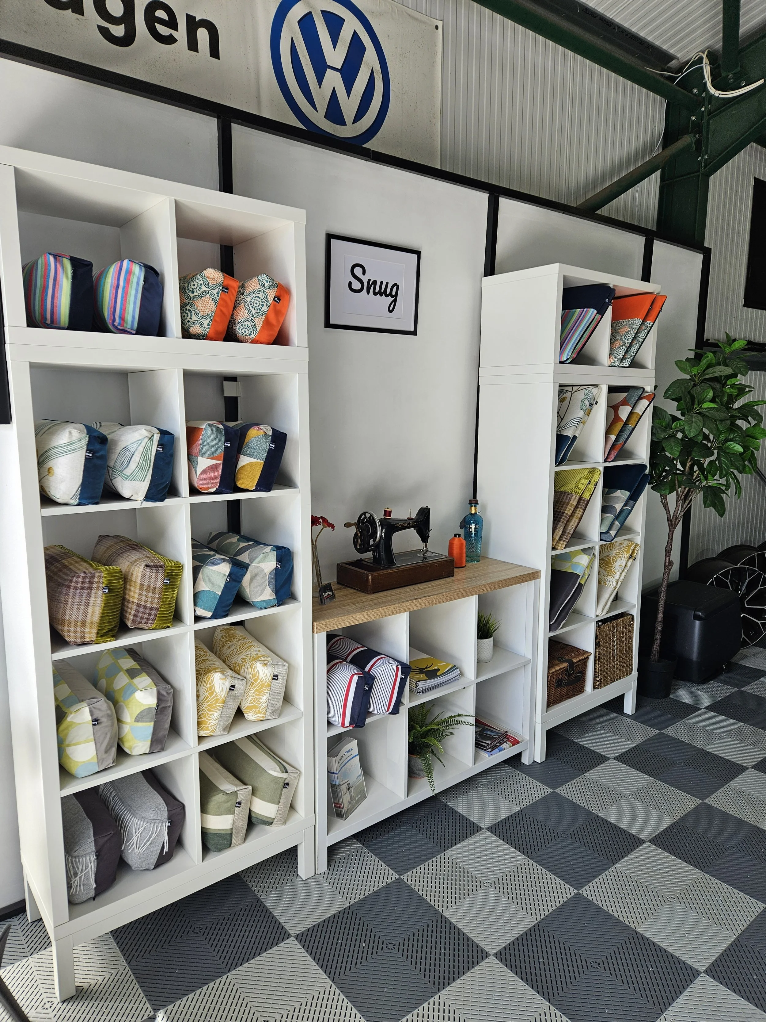 Display of folded fabric bags on white shelving units in a store, with a sign reading 'Snug' on the wall behind, in a space with checkered black and gray flooring and a large window or wall sign with a Volkswagen logo.