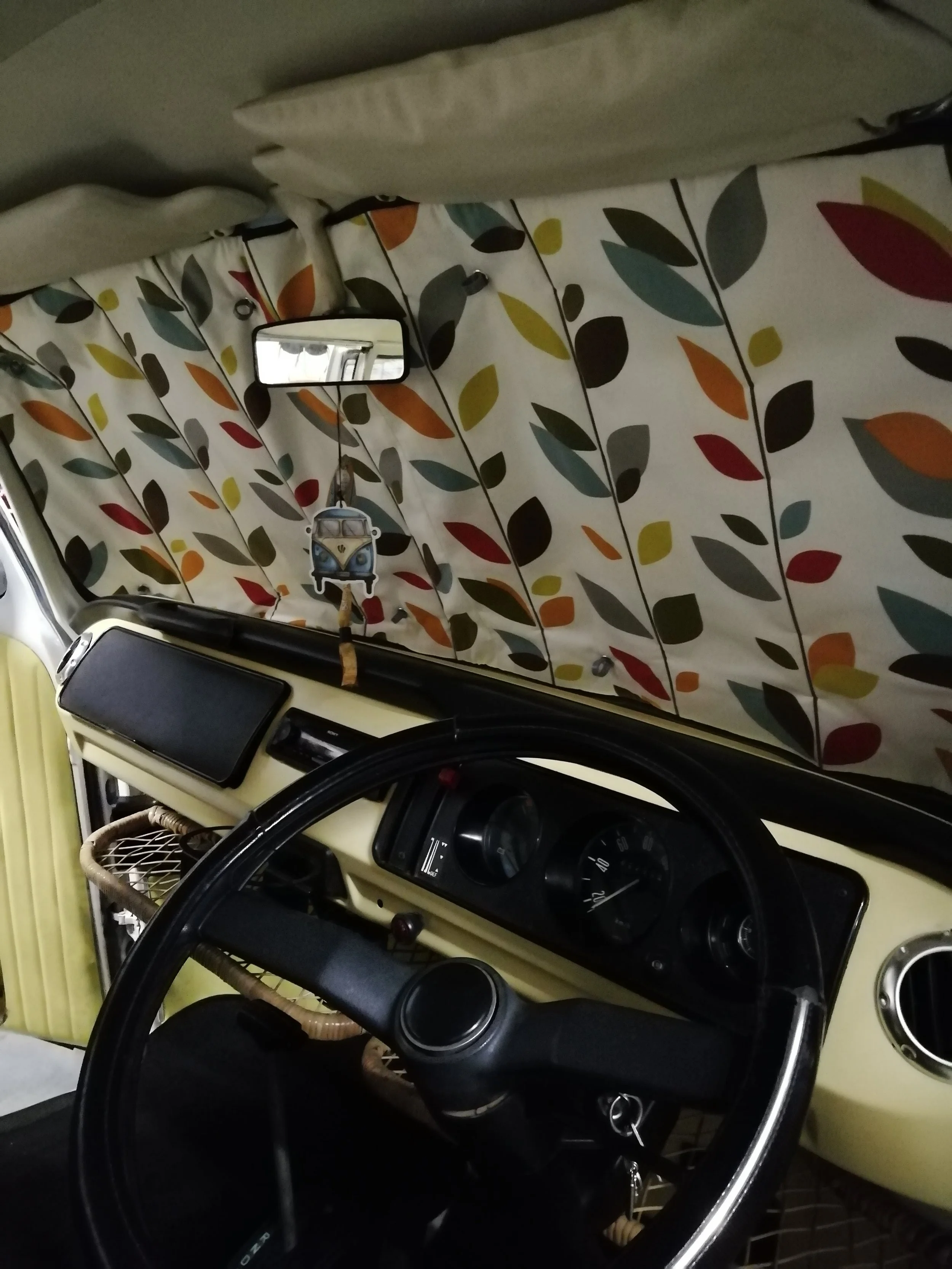 Interior of a vintage Volkswagen van with a black steering wheel, dashboard, a small side mirror, and a curtain with a colorful leaf pattern.