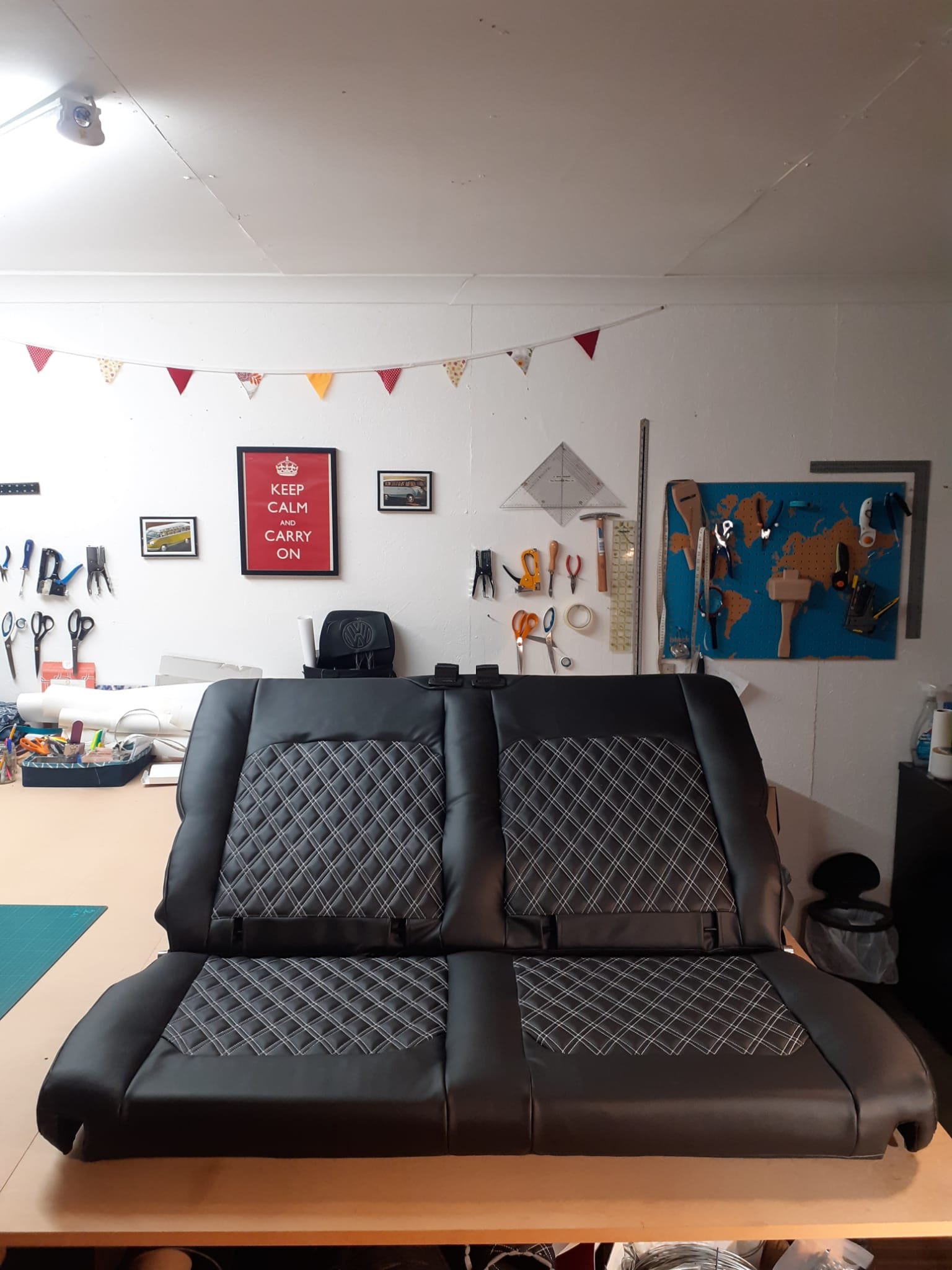 A workroom with a black leather couch facing forward, a wall decorated with framed pictures, a string of triangular colorful flags hanging from the ceiling, and various tools and supplies on a pegboard and shelf.