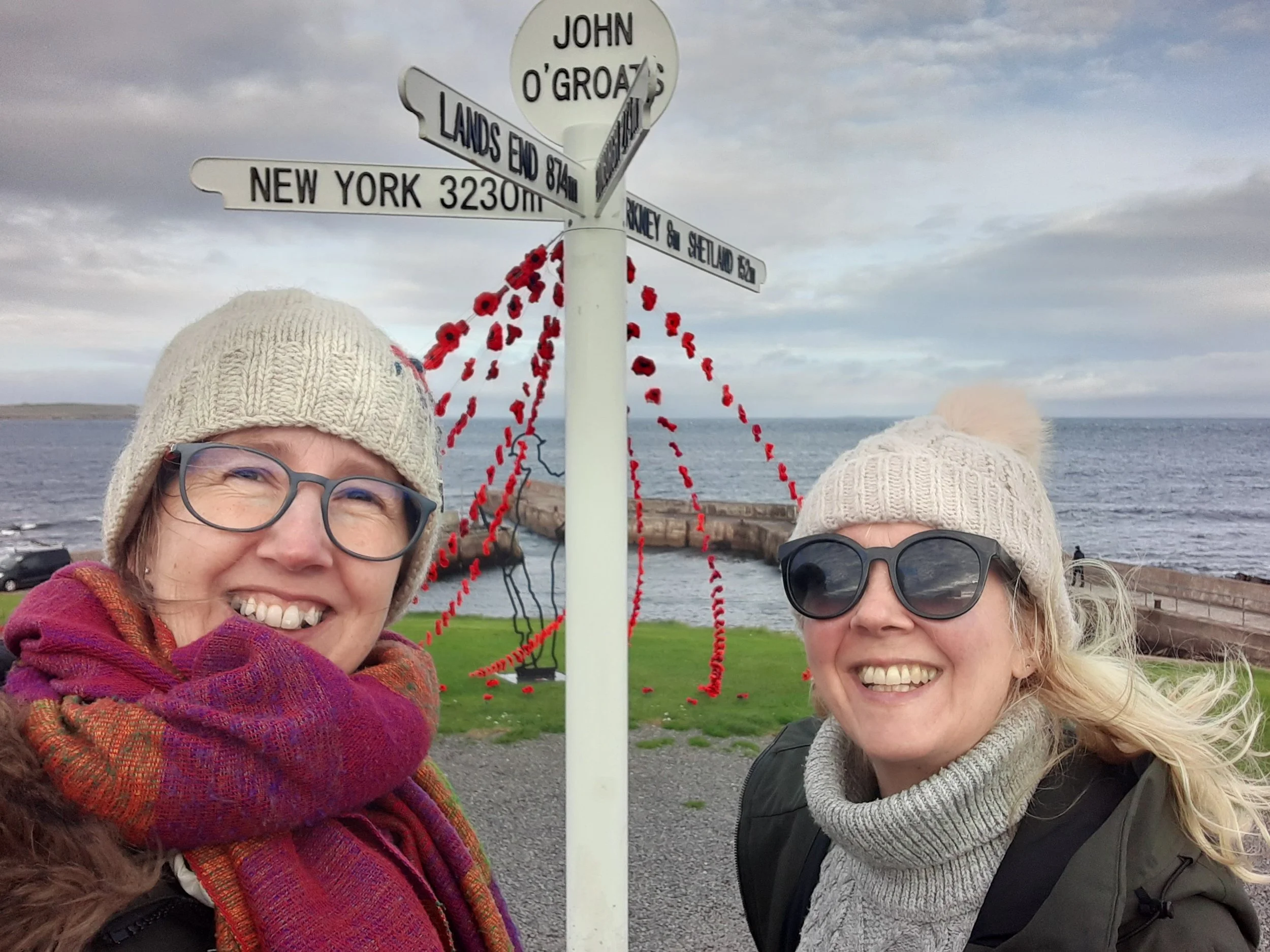 Two women standing outdoors near a seaside location, smiling for a selfie. They are wearing winter hats and scarves. Behind them is a signpost with multiple directional signs, some reading 'John O'Groats,' 'Land's End,' 'New York,' and 'Mickey & Setland.' There are red poppy flowers attached to the signpost, and the sky is cloudy over the ocean.