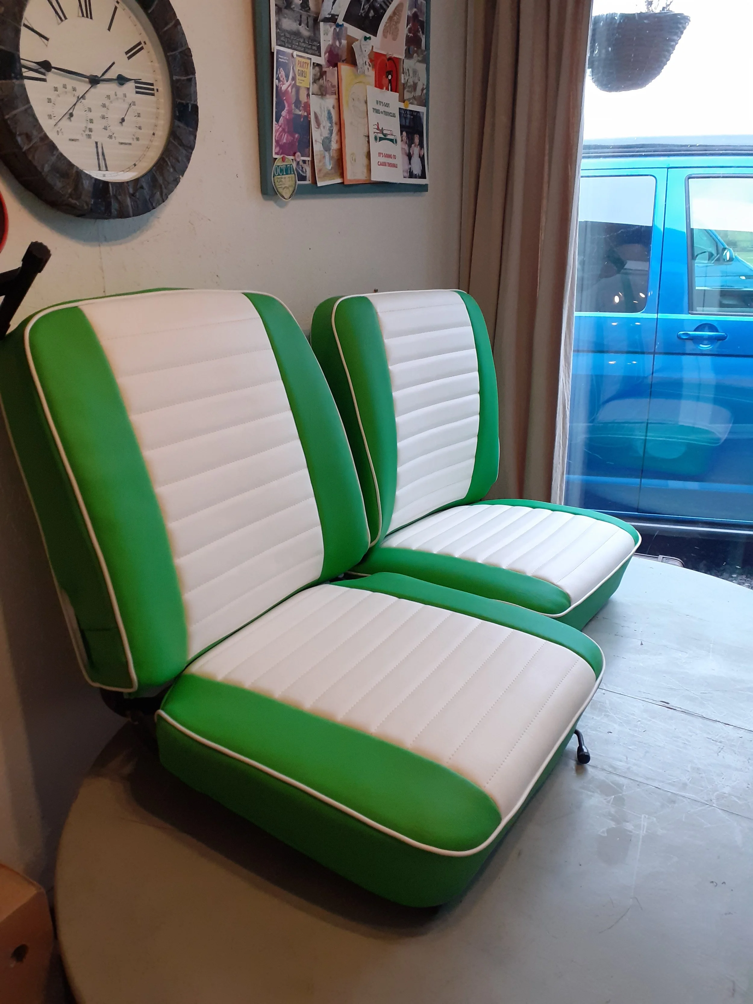 Retro green and white diner-style chairs in a room with a wall clock, a corkboard with photos and notes, and a large window with curtains overlooking a blue vehicle outside.