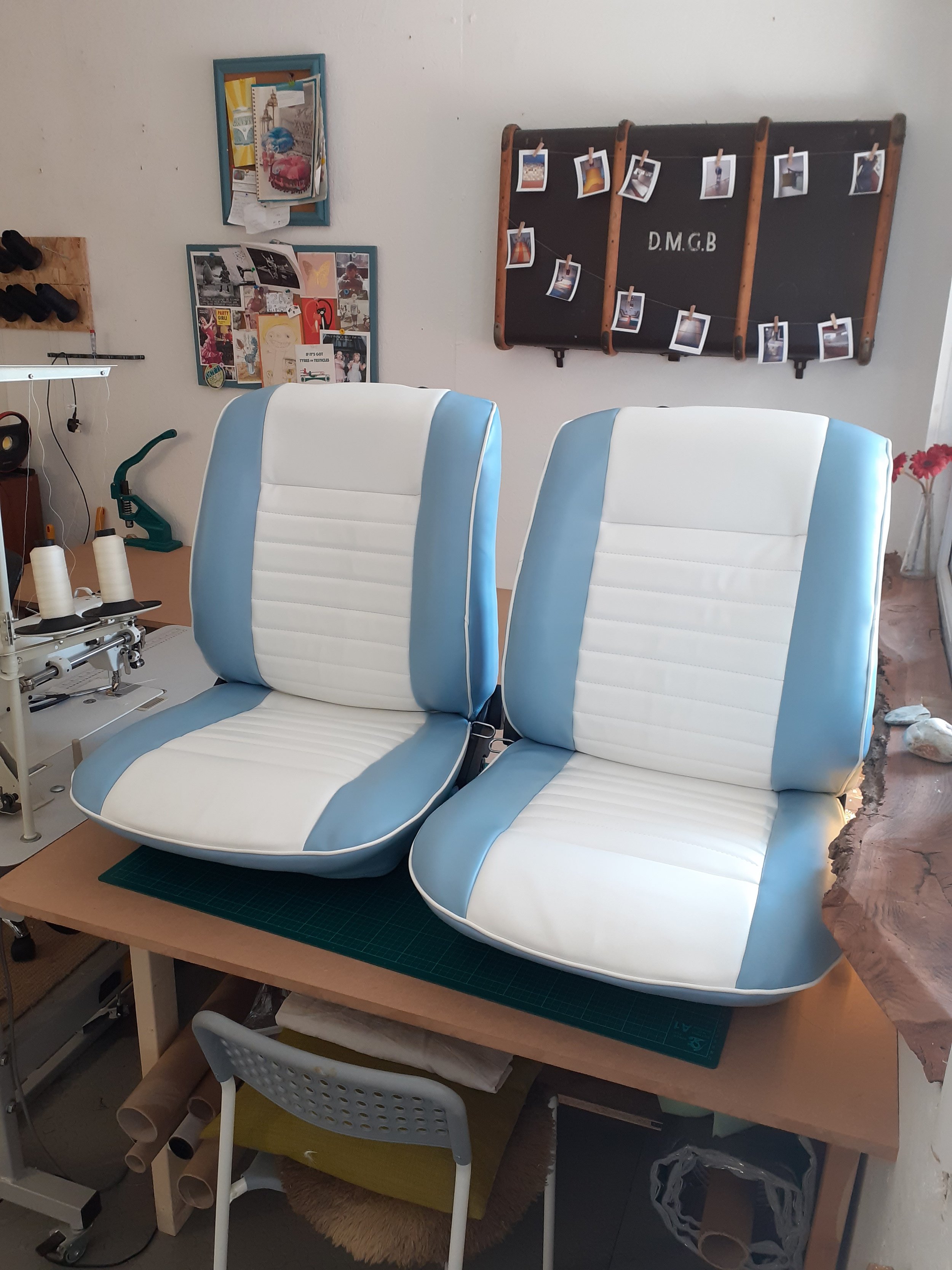 A work station with two white and blue padded chairs placed in front of a table. There is a green cutting mat on the table, and sewing-related items, including rolls of fabric and a sewing machine, underneath. Behind, there is a wall with a bulletin 