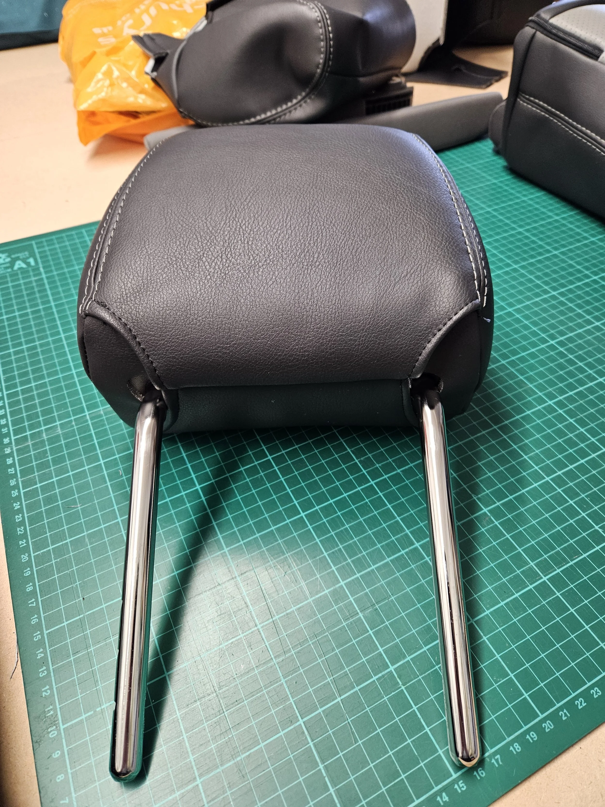 Close-up of a black leather motorcycle seat with metallic rails, placed on a green cutting mat. In the background, there is a yellow plastic bag, a black cylindrical object, and other black leather cushions.