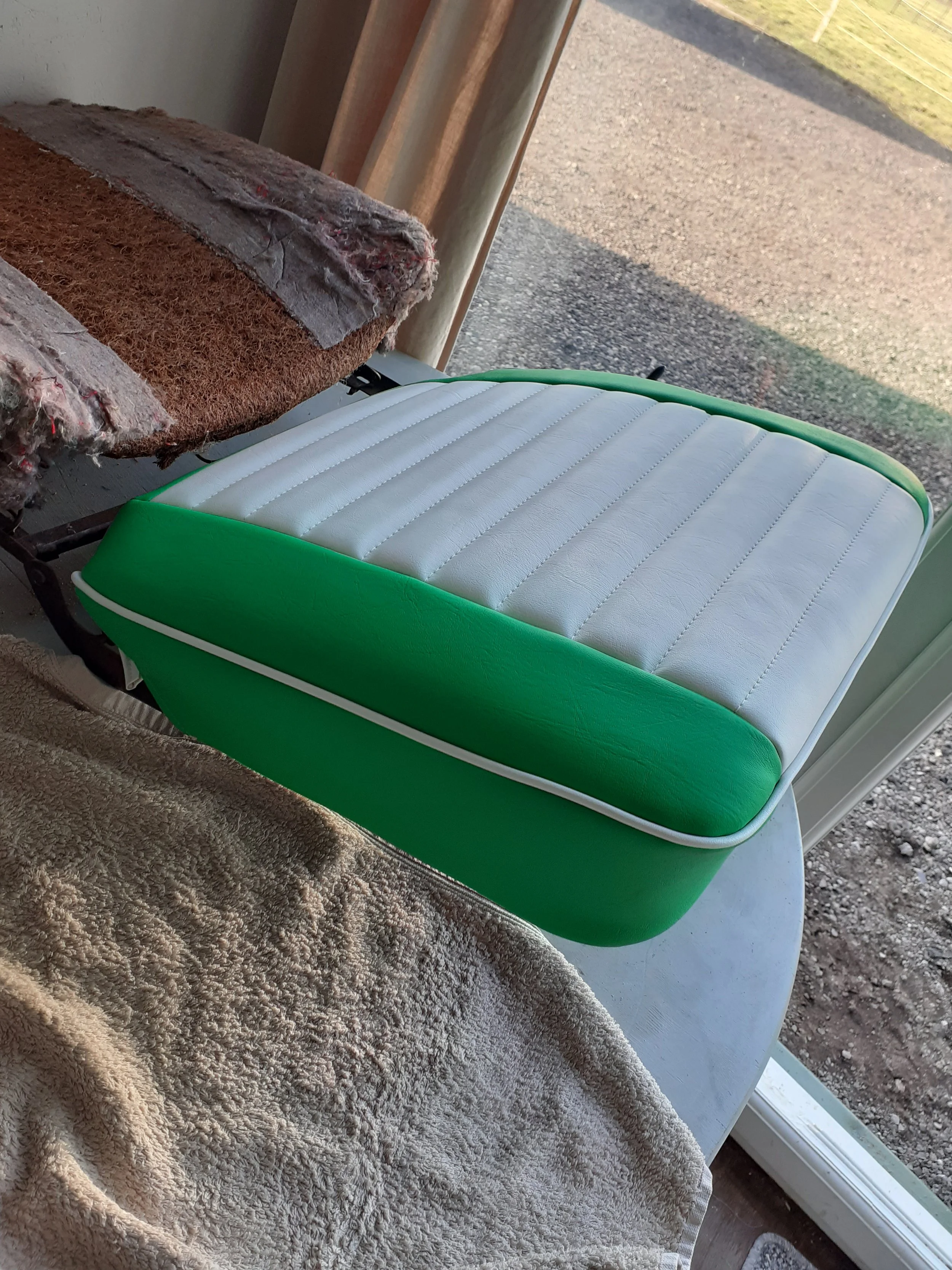 A table with a textured beige towel, a vintage worn-out chair with fabric showing signs of wear, and a modern green and white padded chair on a porch with a gravel path visible outside.
