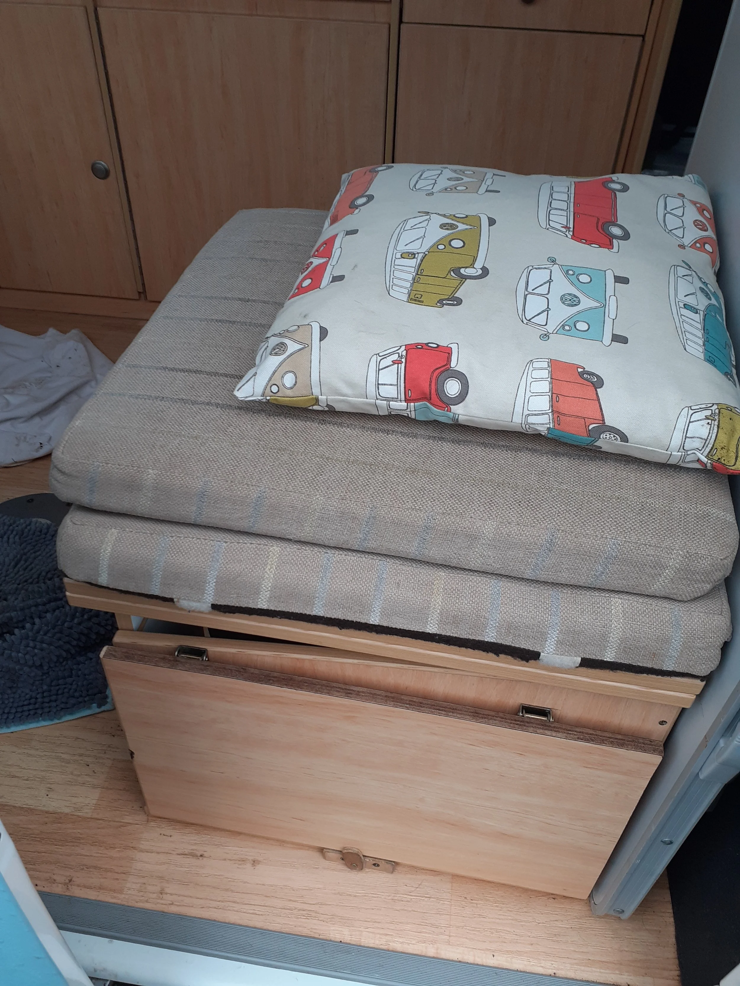 A folded beige cushioned seat with a drawer underneath, placed next to a wooden cabinet with vertical grain, and a bed with a pillow that has a white cover with colorful vintage vehicle illustrations.