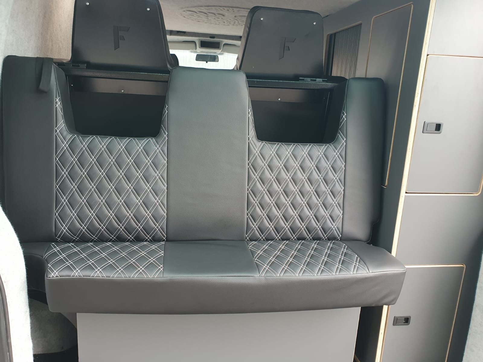 Black and gray quilted bench seat with two black headrests in the interior of a vehicle, likely a camper or van, with storage cabinets nearby.