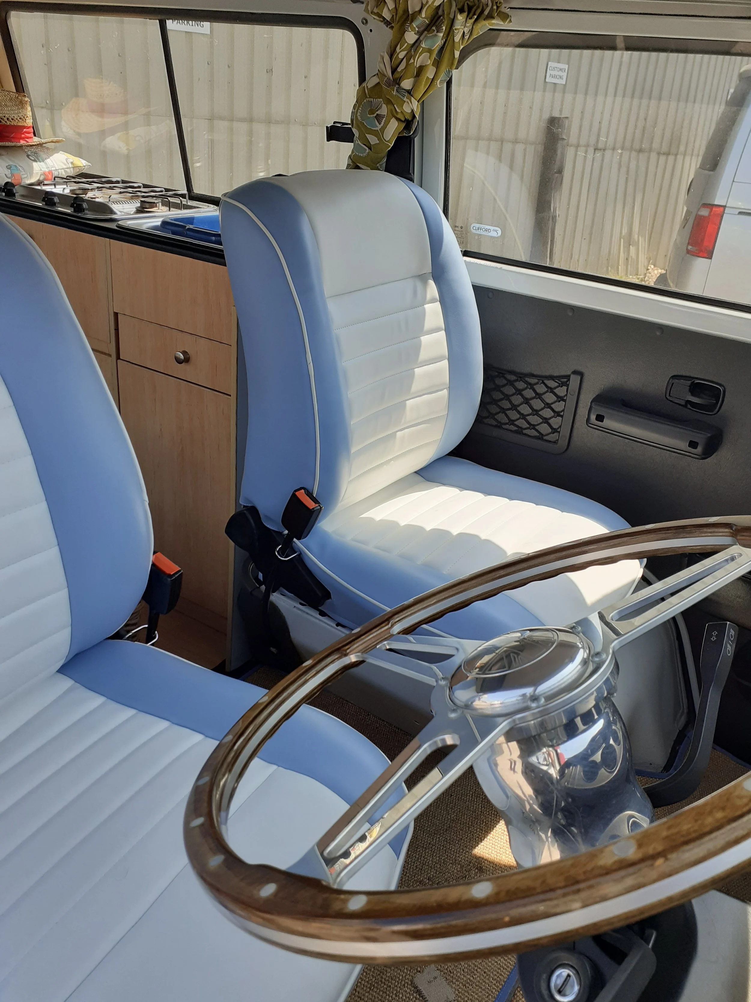 Interior of a vintage vehicle with two front seats in white and blue upholstery, a wooden steering wheel, a small wooden cabinet, and a large window showing parked cars and a wooden fence outside.