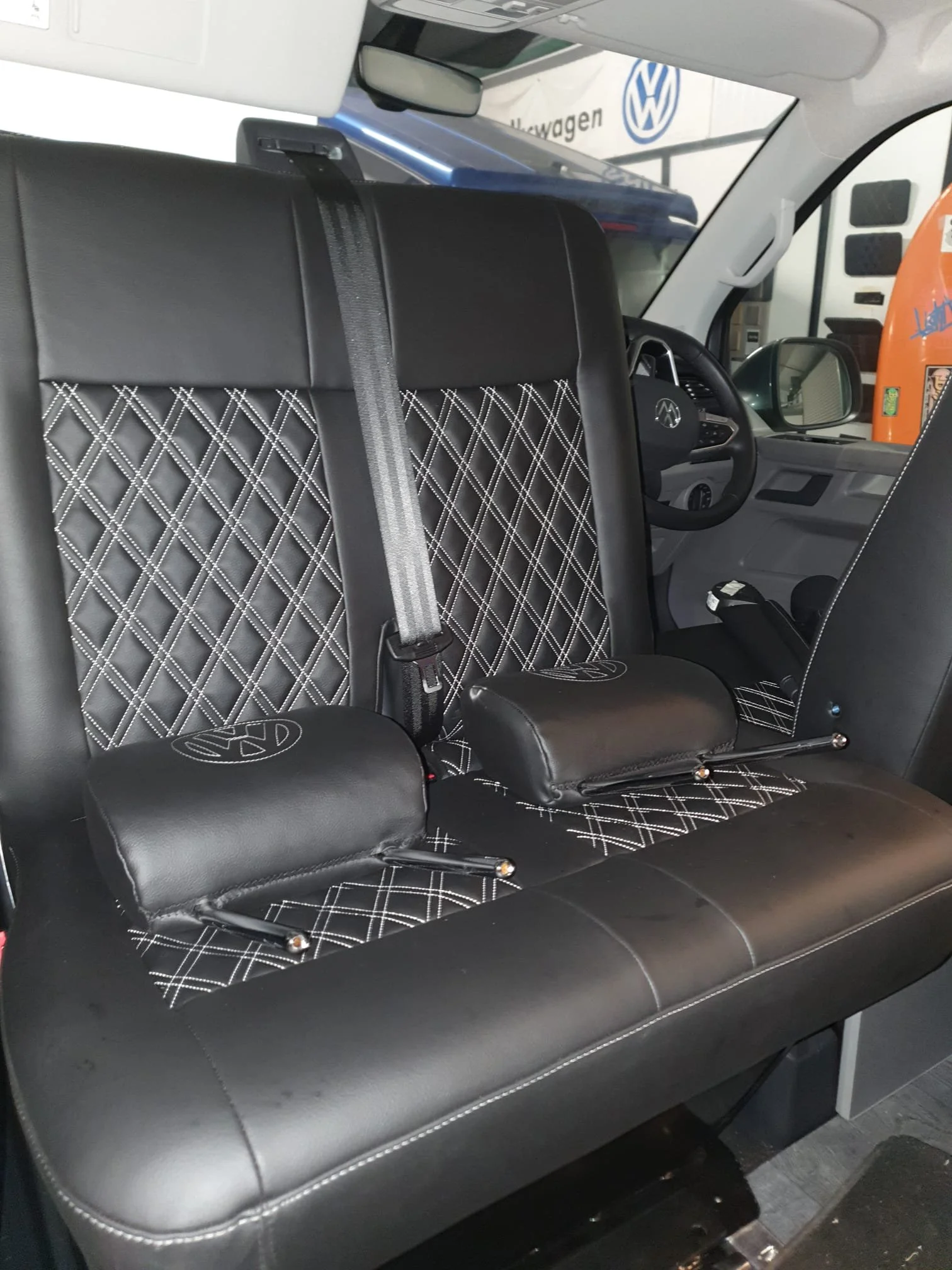 The interior of a vehicle showing a black leather seat with diamond-patterned stitching, a black seatbelt, and a headrest with the VW logo. The dashboard and steering wheel are visible, with a VW emblem on the steering wheel. In the background, part 