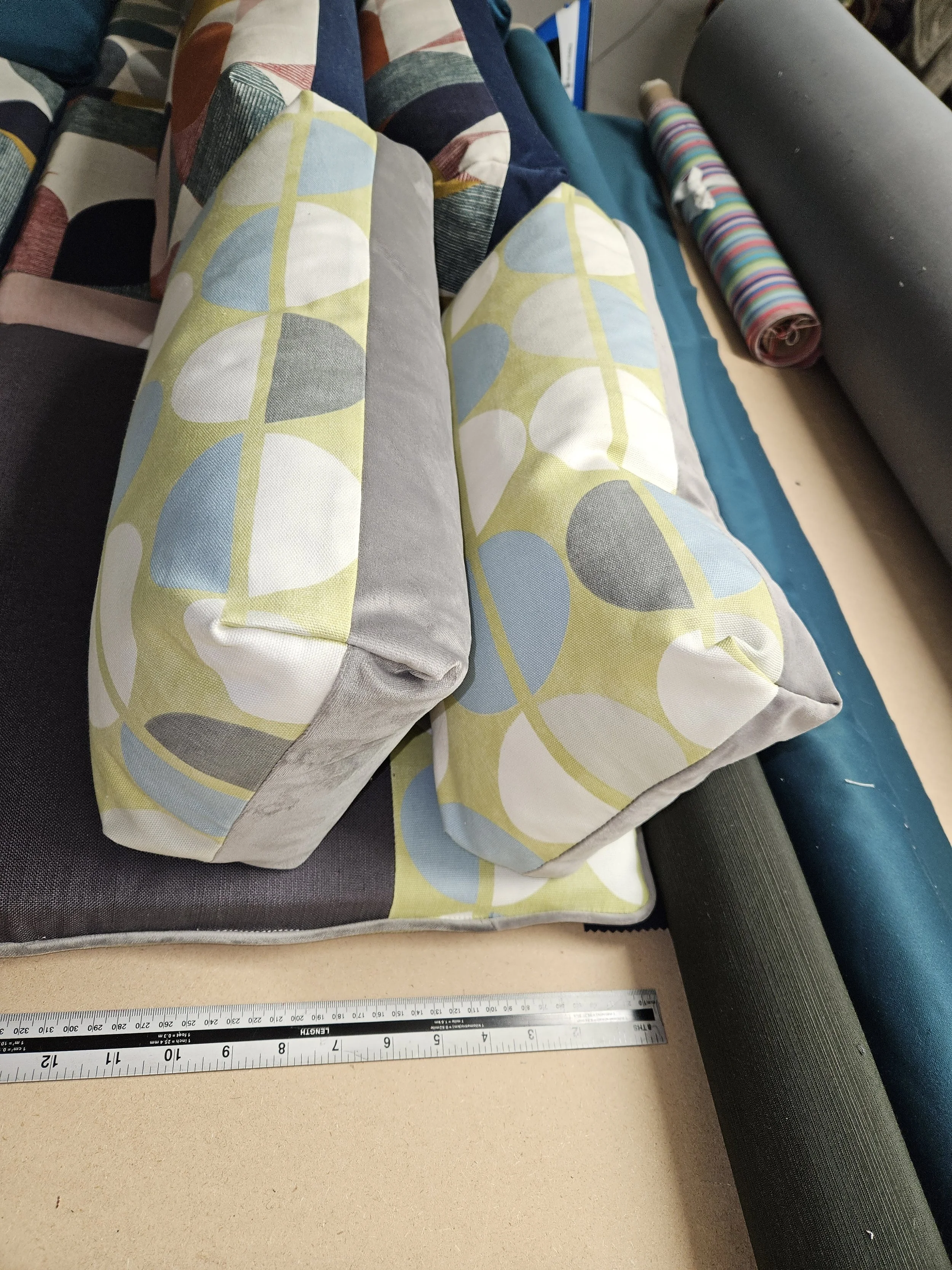 Fabric rolls on a table, including a roll with a geometric pattern in mint green, light blue, white, gray, and yellow. There is a ruler at the bottom of the image and additional fabric rolls in various colors surrounding the main fabric.