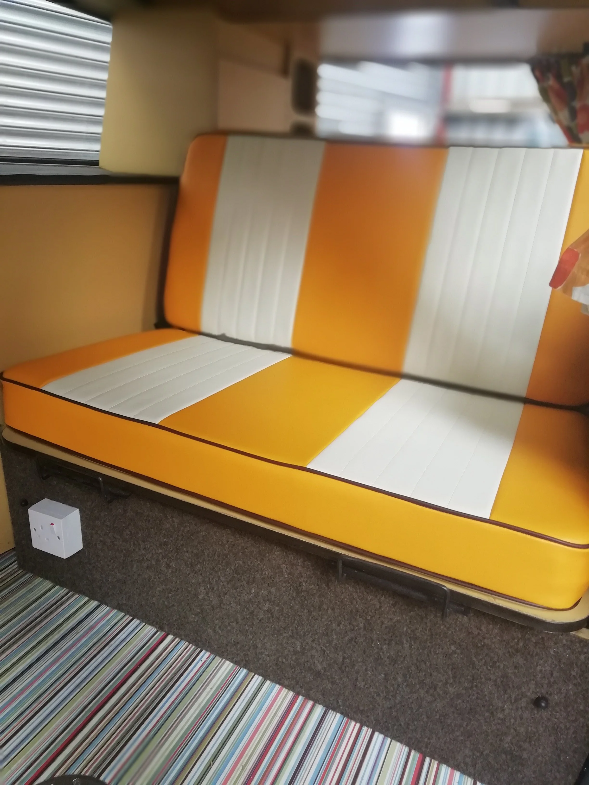 An orange and white striped cushioned chair next to a gray carpeted floor and a multicolored striped rug, with part of a window and a white power strip visible.