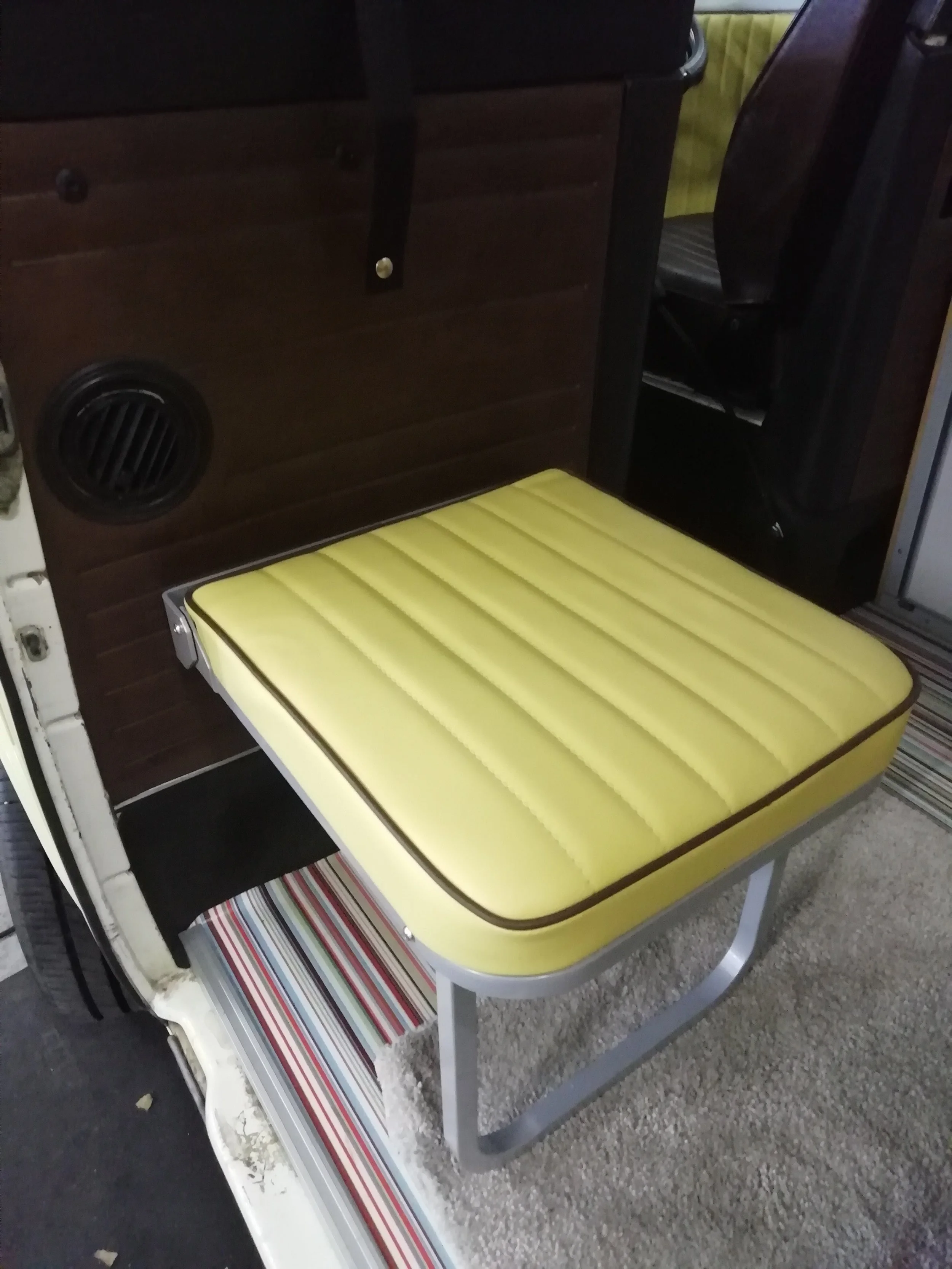 A yellow mattress with brown borders on a gray metal bed frame in front of a plastic shelf with striped material. A brown wooden dresser with a circular vent and black elastic strap is behind the bed.