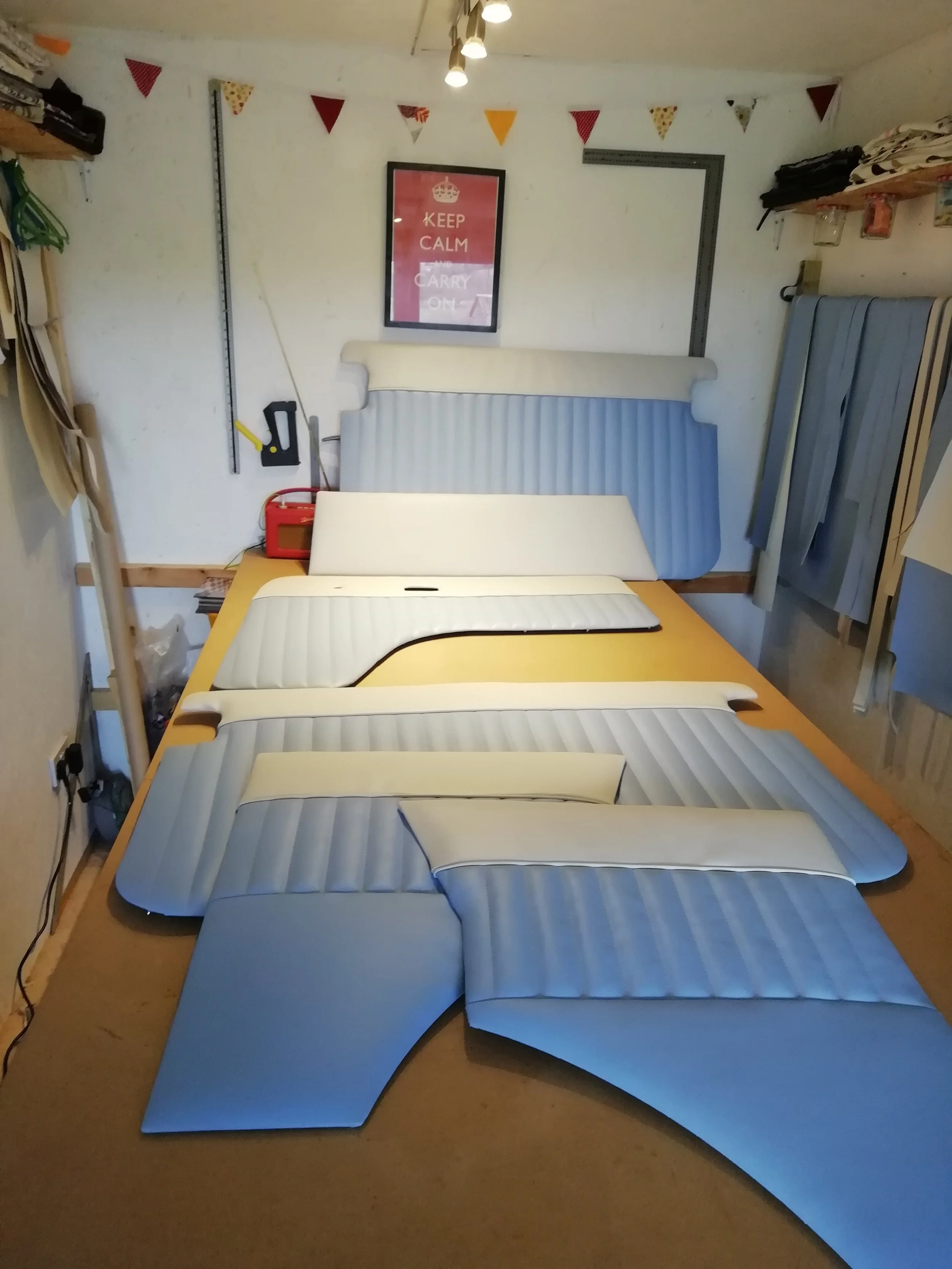 A cluttered room with disassembled beds, a wooden table with mattress parts, shelves with fabric and supplies, a framed 'Keep Calm and Carry On' poster, and an orange alarm clock.