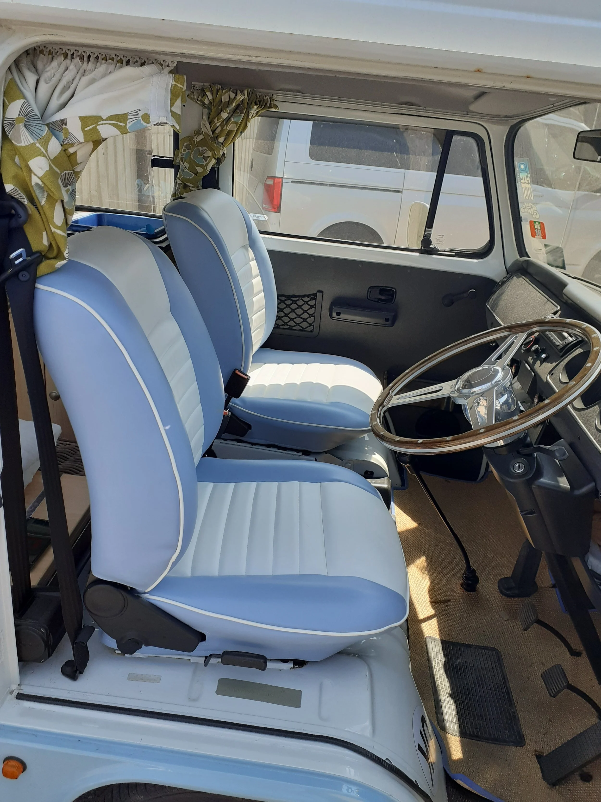 Interior of a vintage van showing white and blue upholstered seats, a wooden steering wheel, vehicle dashboard, pedals, and a large windshield with a view of parked vehicles outside.