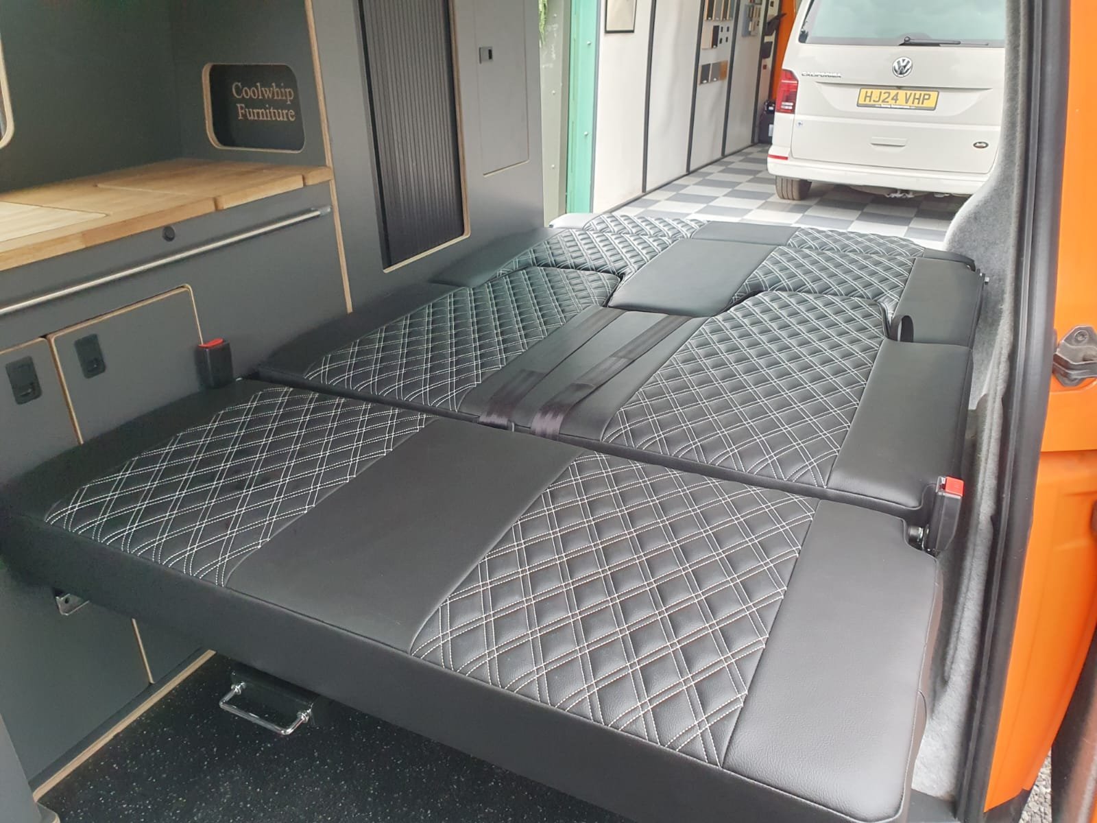 Folded bed inside a camper van with black quilted mattress, black seat belts, interior cabinets, and a view of a white van and checkered floor outside.