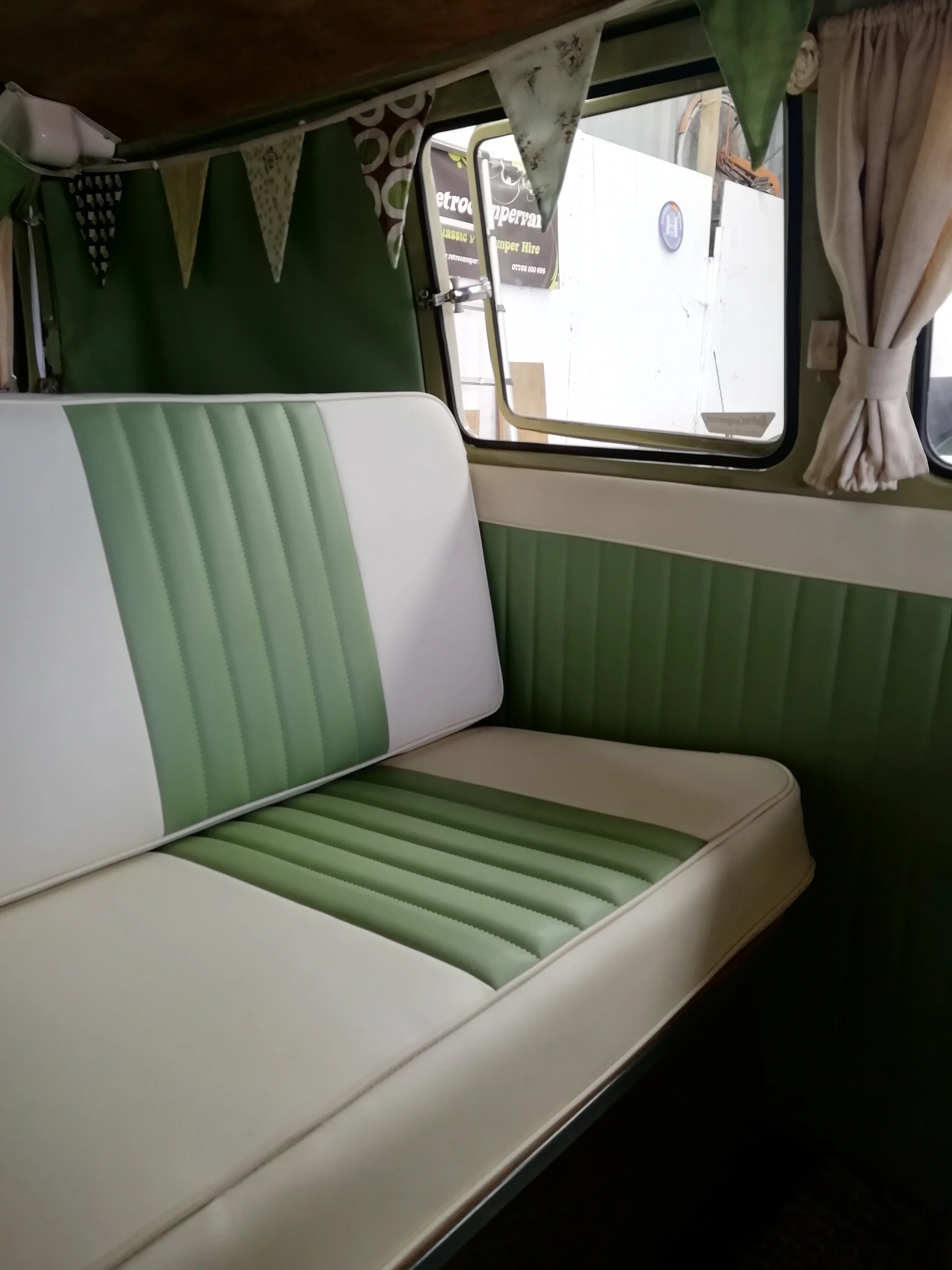 Inside of a camper van showing a green and white striped upholstered seating area, with colorful bunting hanging above and a window with beige curtains on the right side.