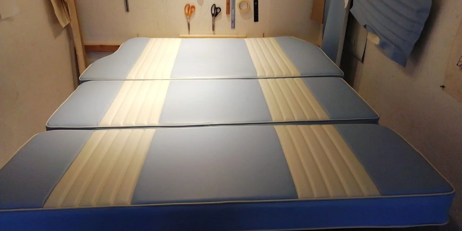 A room with a bed with three mattresses stacked together, a wall with hanging tools including scissors, a ruler, and tape, and a hanging blue quilt or blanket.
