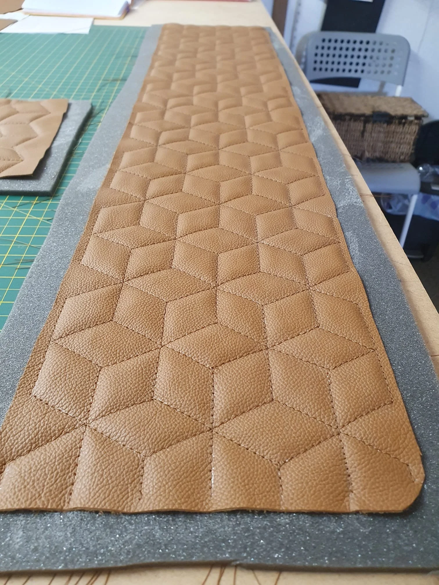 Close-up of a long strip of tan quilted leather with a hexagonal pattern, placed on a worktable covered with a green cutting mat, with some papers and tools in the background.