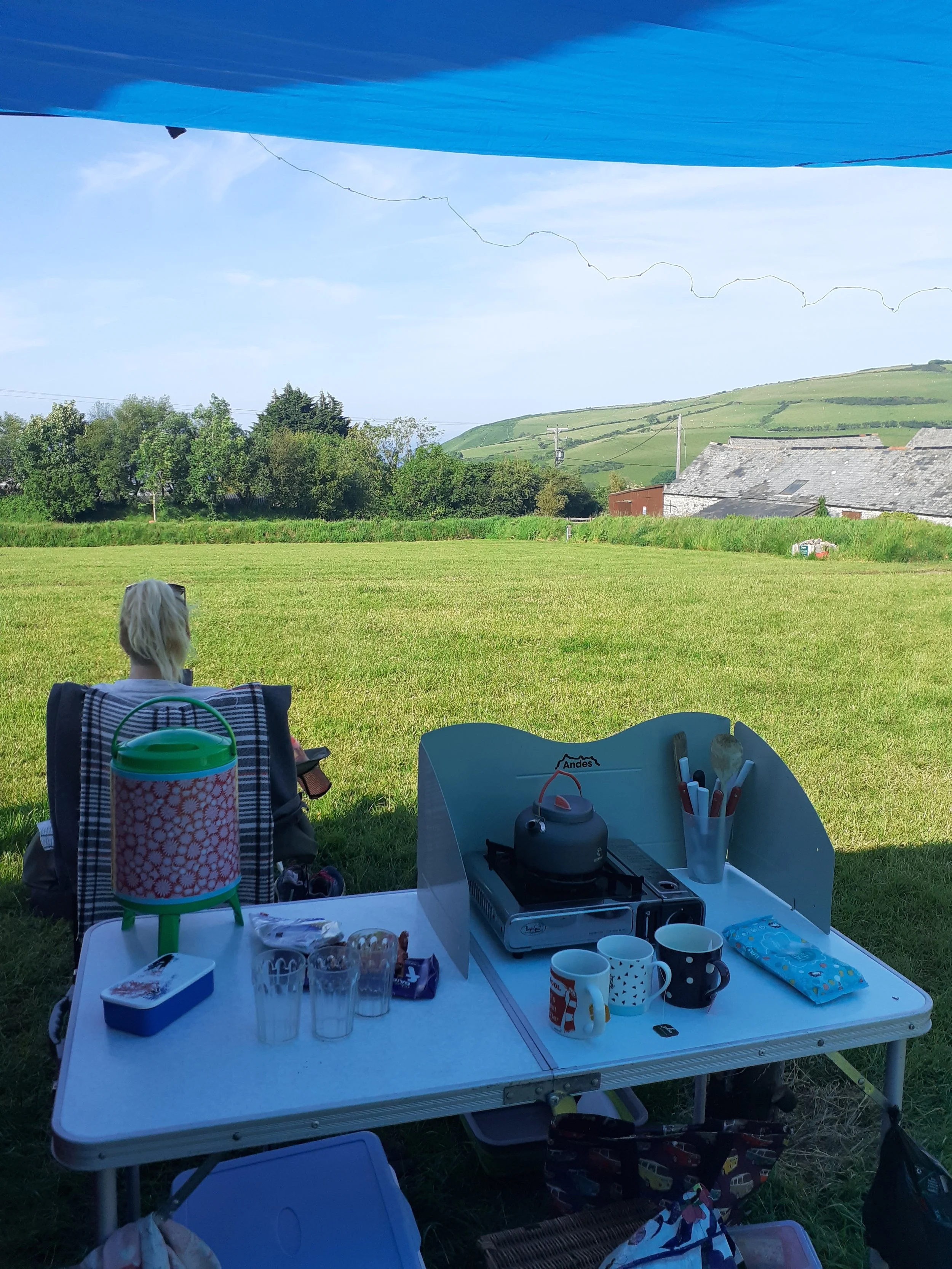 A table set up outdoors with cups, plates, and a portable stove, with a scenic green landscape and houses in the background under a blue sky.