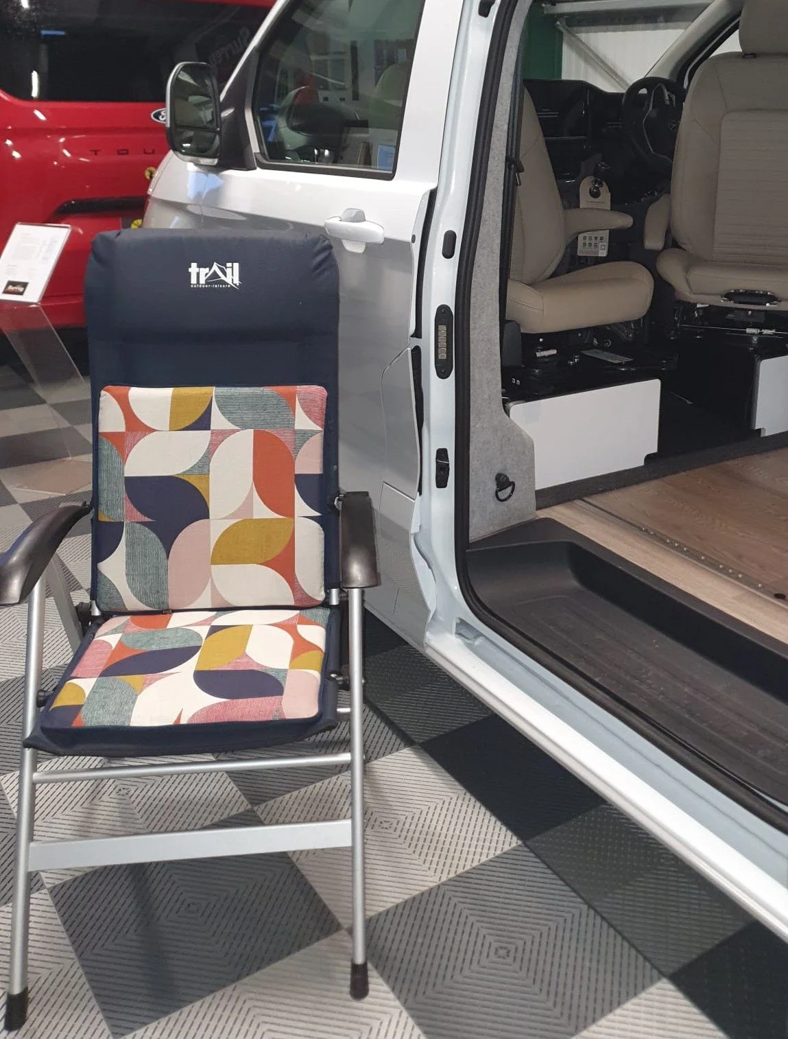A wheelchair with a colorful geometric patterned cushion positioned next to a white van with its doors open, revealing an interior with beige seats inside a dealership or exhibition space.