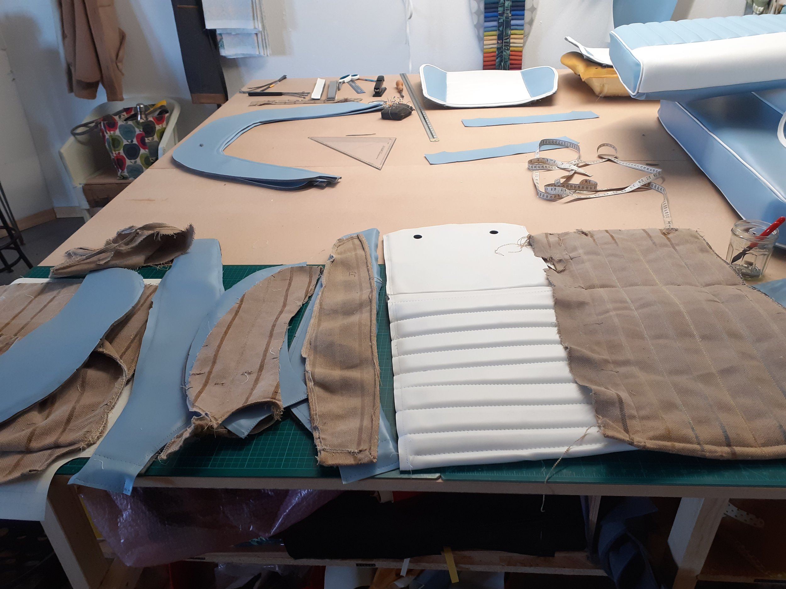 A workspace with fabric pieces, blue patterns, measuring tape, rulers, and cutting tools on a wooden table. It appears to be for sewing or upholstery projects.