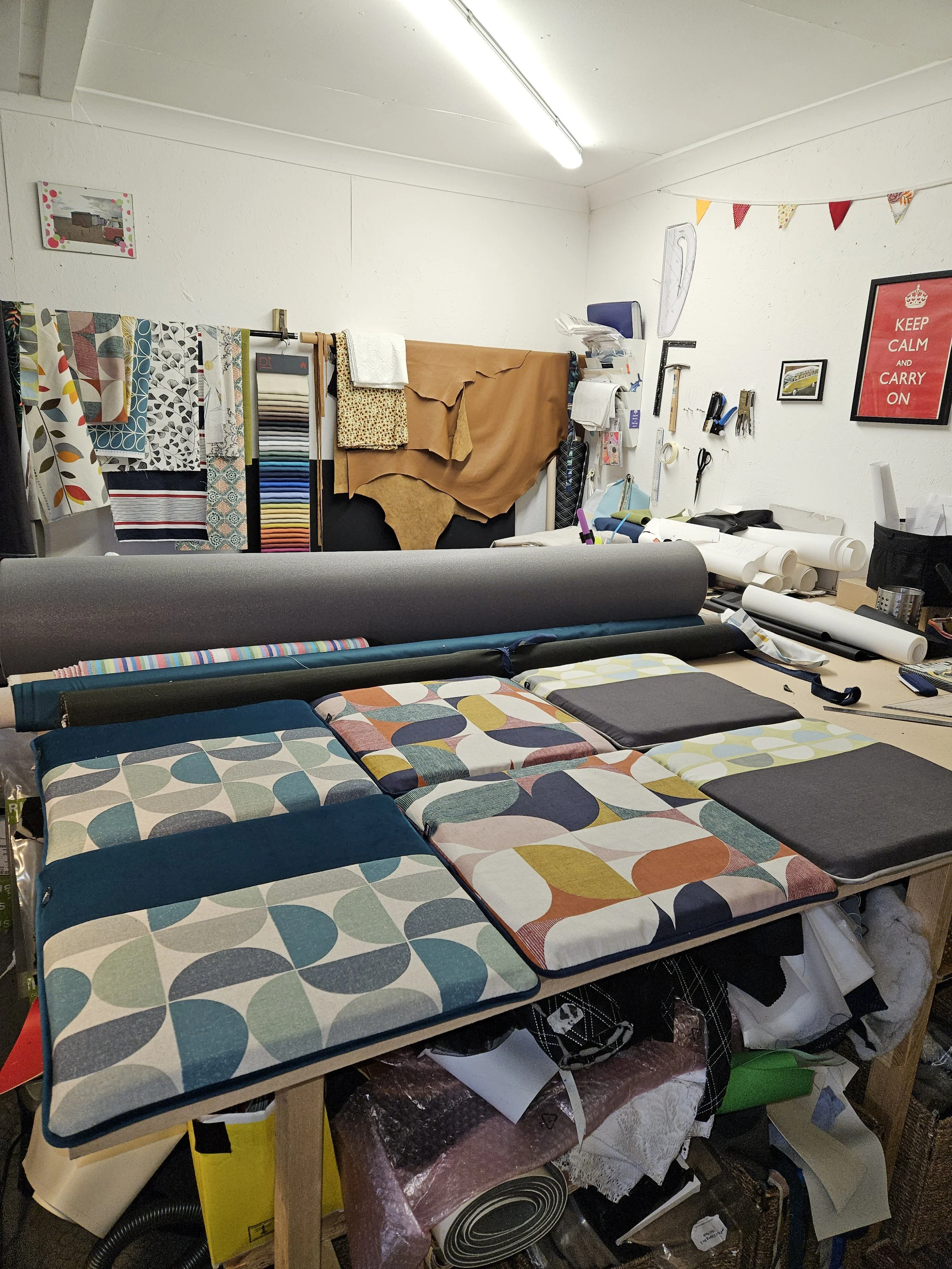 A workspace with fabric swatches, rolls of material, and tools. There are fabric samples hanging on a rack, and rolls of fabric and wallpaper on the table. The wall has various tools, scissors, and a framed sign that says 'Keep Calm and Carry On.'