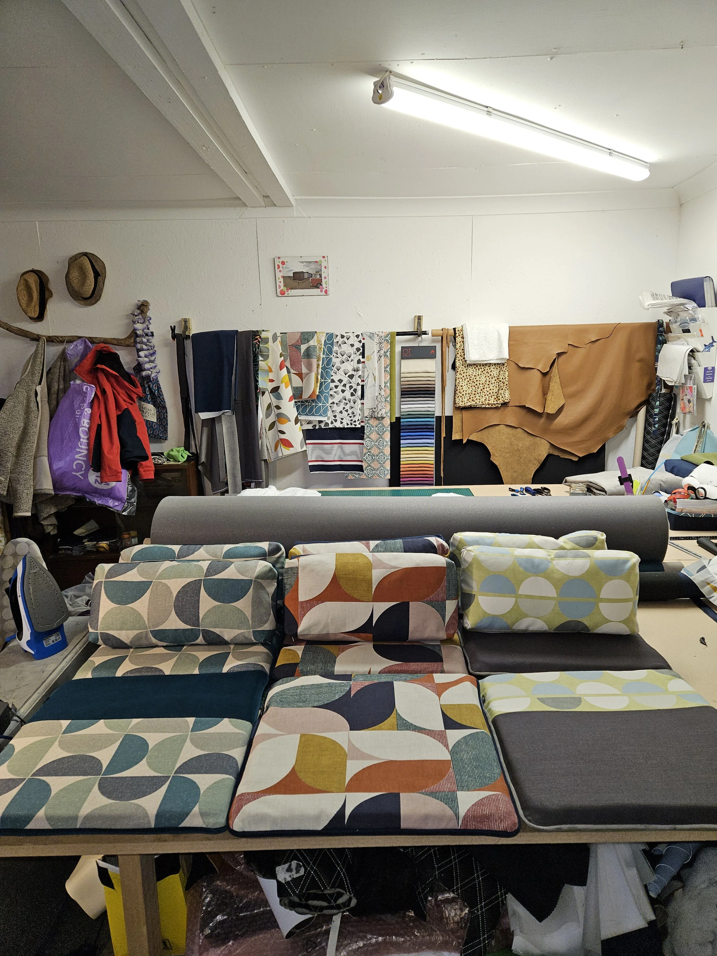 A workspace with fabric swatches and sewing supplies. The foreground shows fabric-covered cushions with various patterns. The background has hanging textiles, a small framed picture on the wall, and items like hats and bags hanging on the wall.