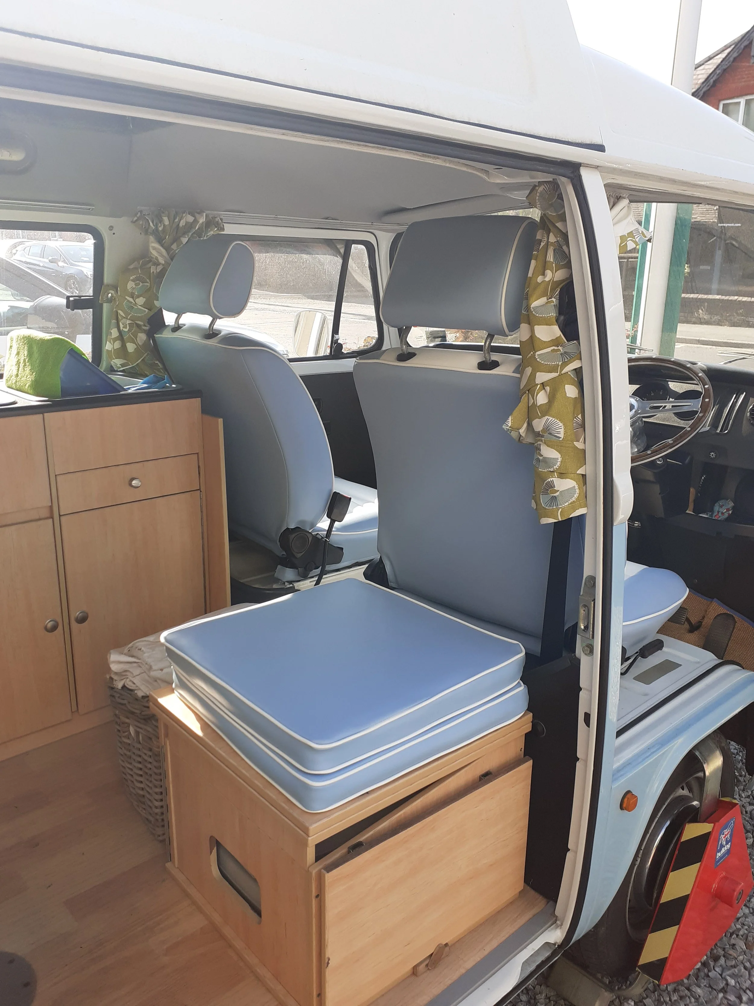 The interior of a camper van showcasing blue cushioned seats, a wooden cabinet with drawers, and a wicker basket with blue cushions on top. The window curtains are yellow with a floral pattern, and there's a view of parked cars outside.