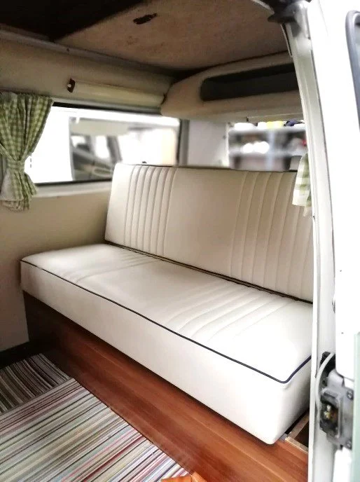 Interior of a vintage camper with a cream-colored bench seat, green checkered curtains, wooden flooring, and a window with a green checkered curtain.