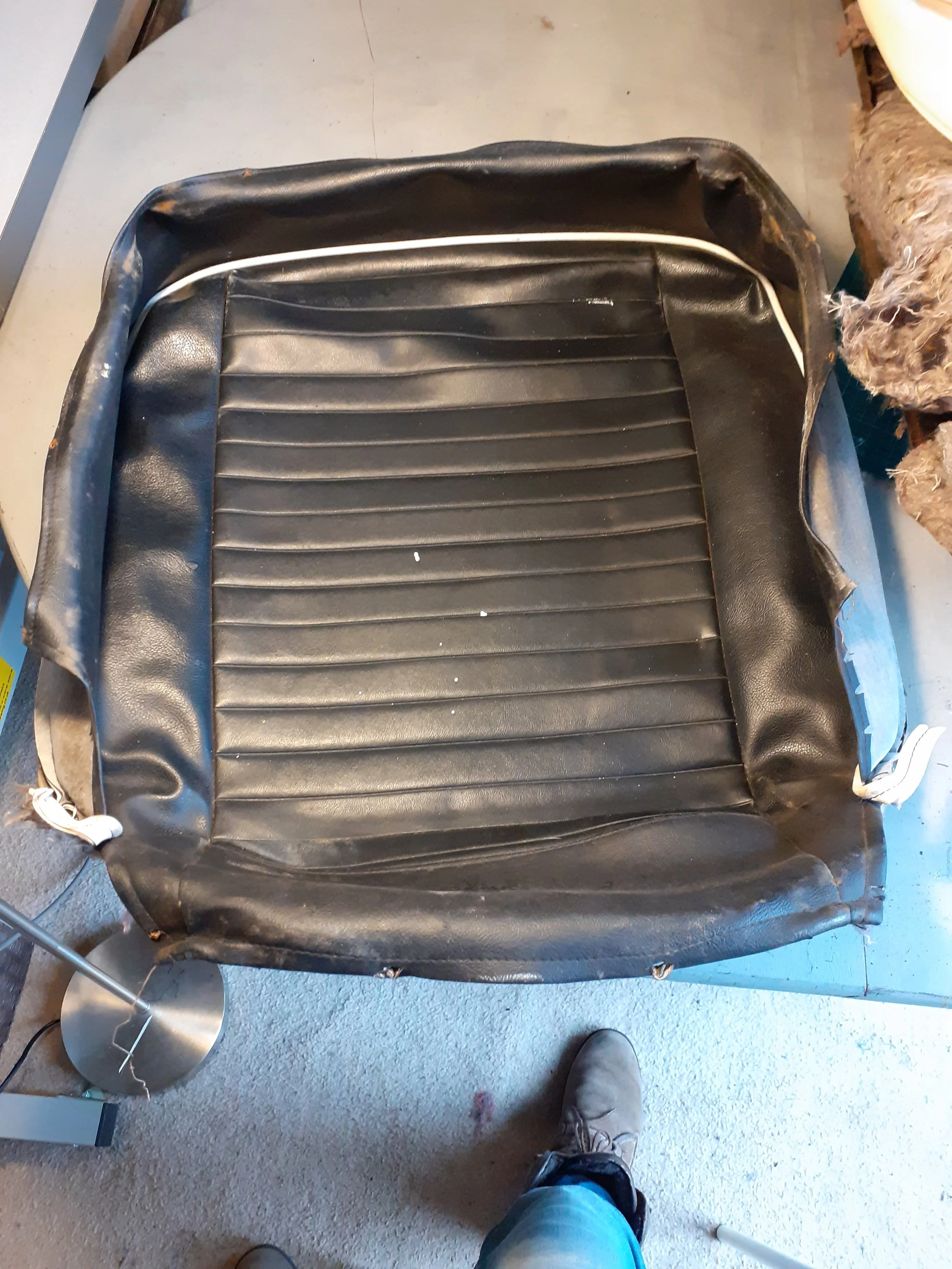 A worn black vinyl car seat with white piping, placed on a floor with a person in jeans and sneakers standing in front of it. To the left, a small metal lamp and other miscellaneous items are partially visible.